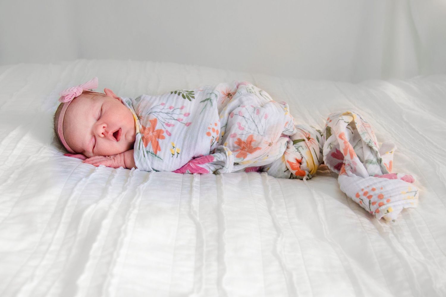 the newborn baby is wrapped in a floral blanket with pinks, orange, yellow and green, wearing a headband bow on a white backdrop