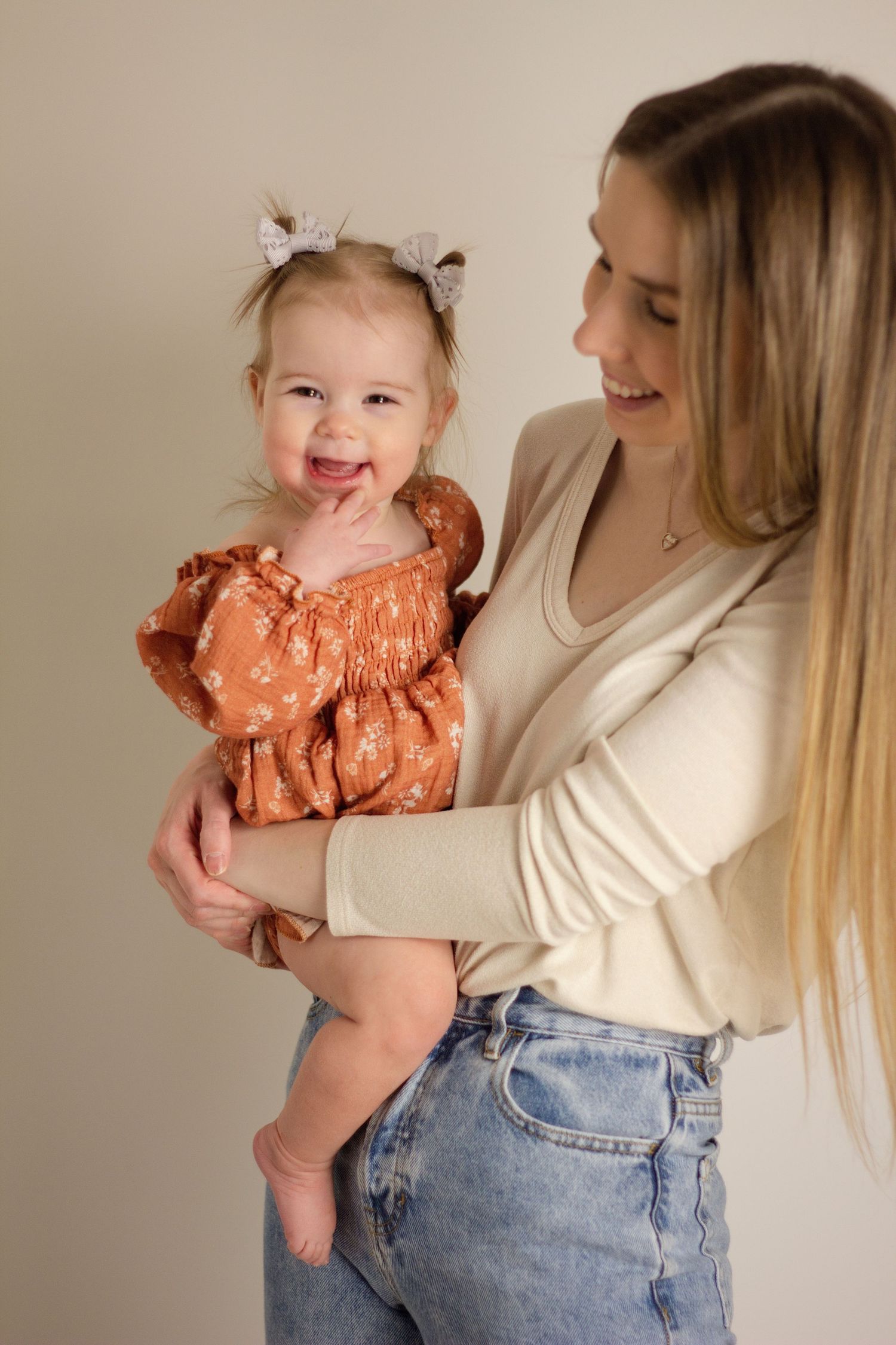 the mom is wearing a beige top and blue jeans holding her one year old daughter in a pink dress with pigtails