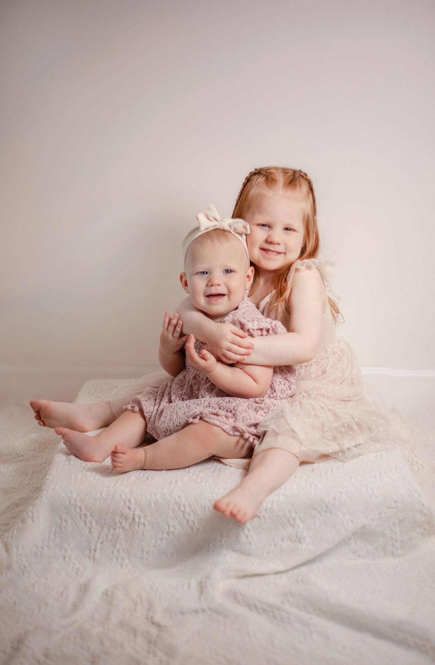 two sisters are sitting with the older ones arms wrapped around, both are wearing shades of light pink, the background is white