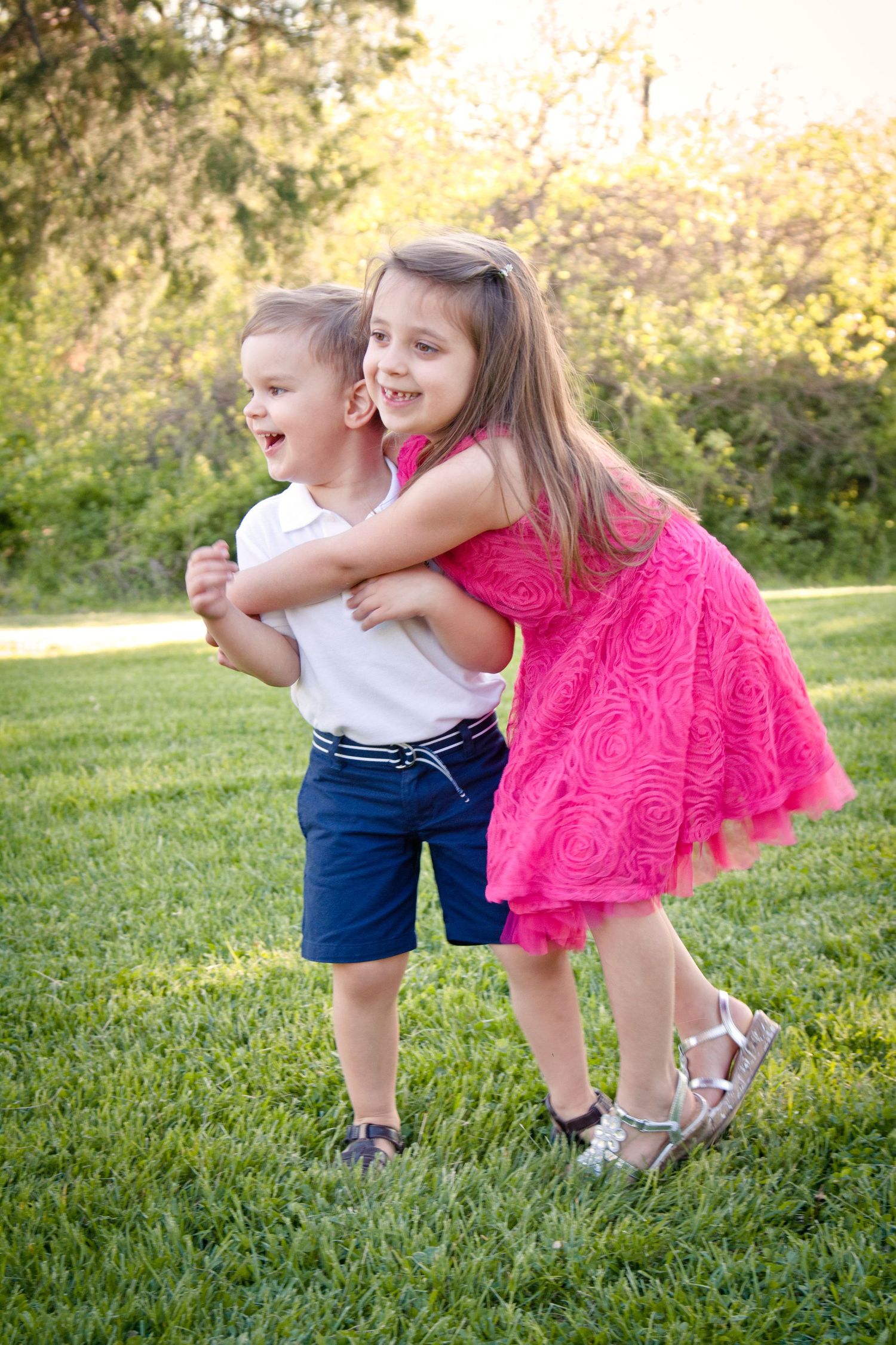 Two children and playing and hugging outdoors, the girl is in a bright pink dress and the little boy is wearing blue shorts and white shirt