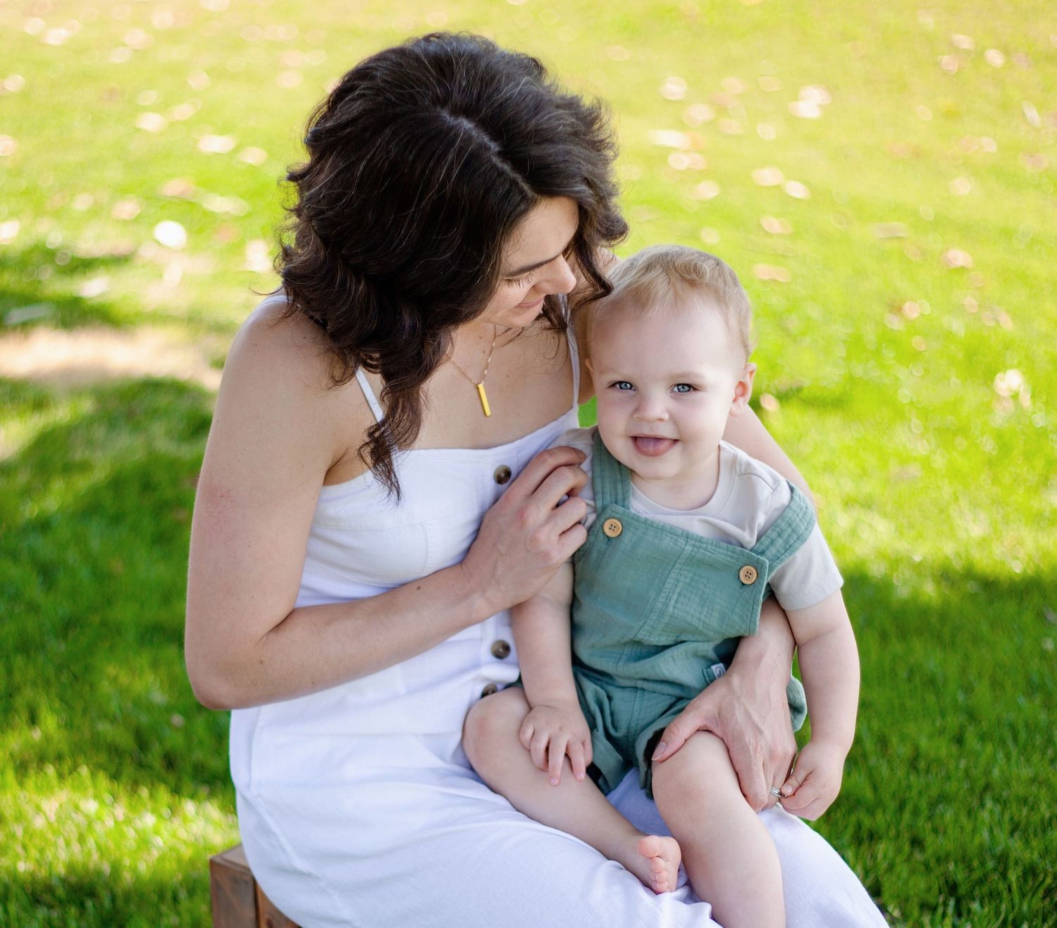 this mommy and me photo shows a mom with dark brown curly hair wearing white and little blonde boy with blue eyes wearing green overalls.