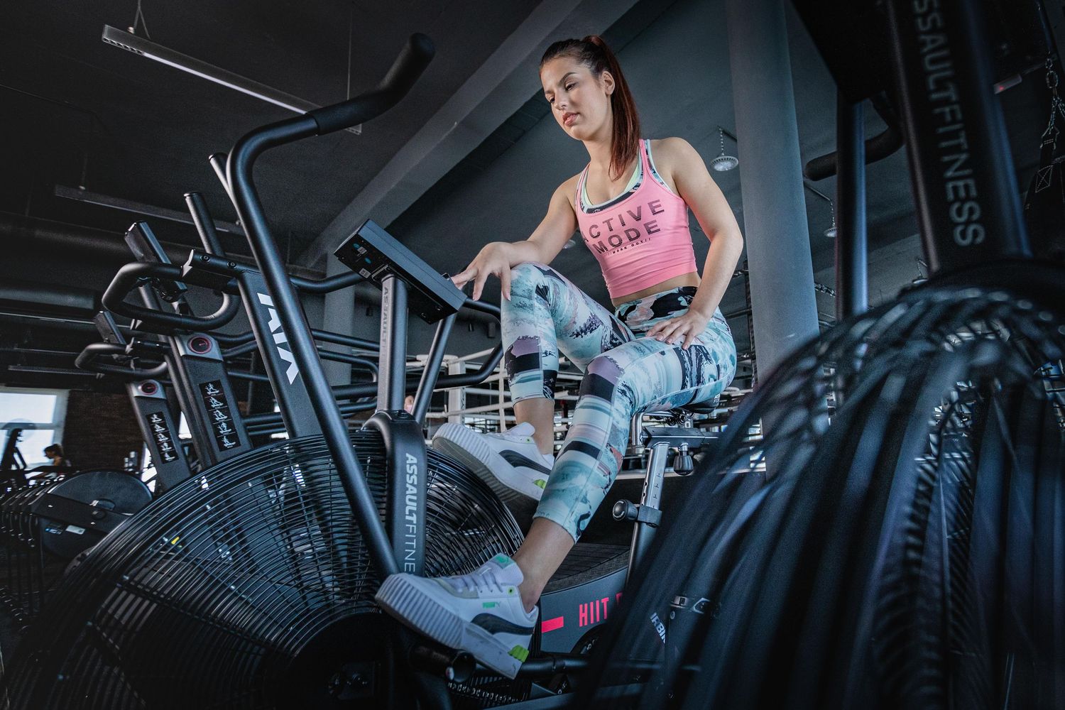 Woman in pink sports top and camo leggings working out on an air bike in a modern gym.