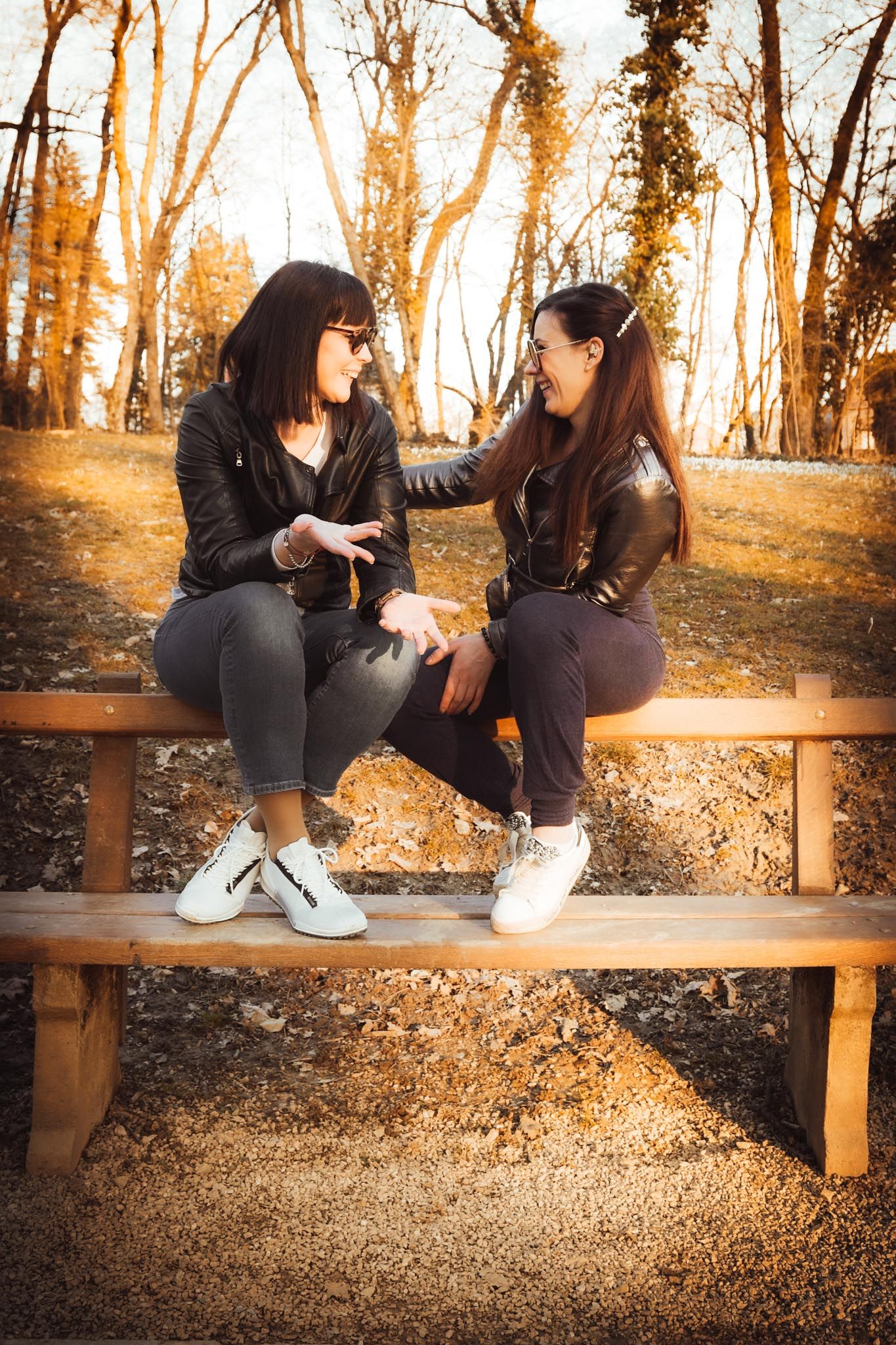 Two women in leather jackets sit on a wooden bench in an autumn park, smiling and chatting in warm golden light.