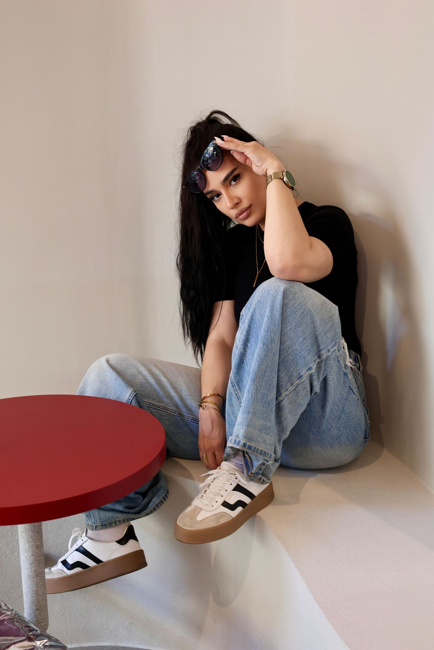 Young woman in jeans and sneakers sitting on floor against white wall next to red table.
