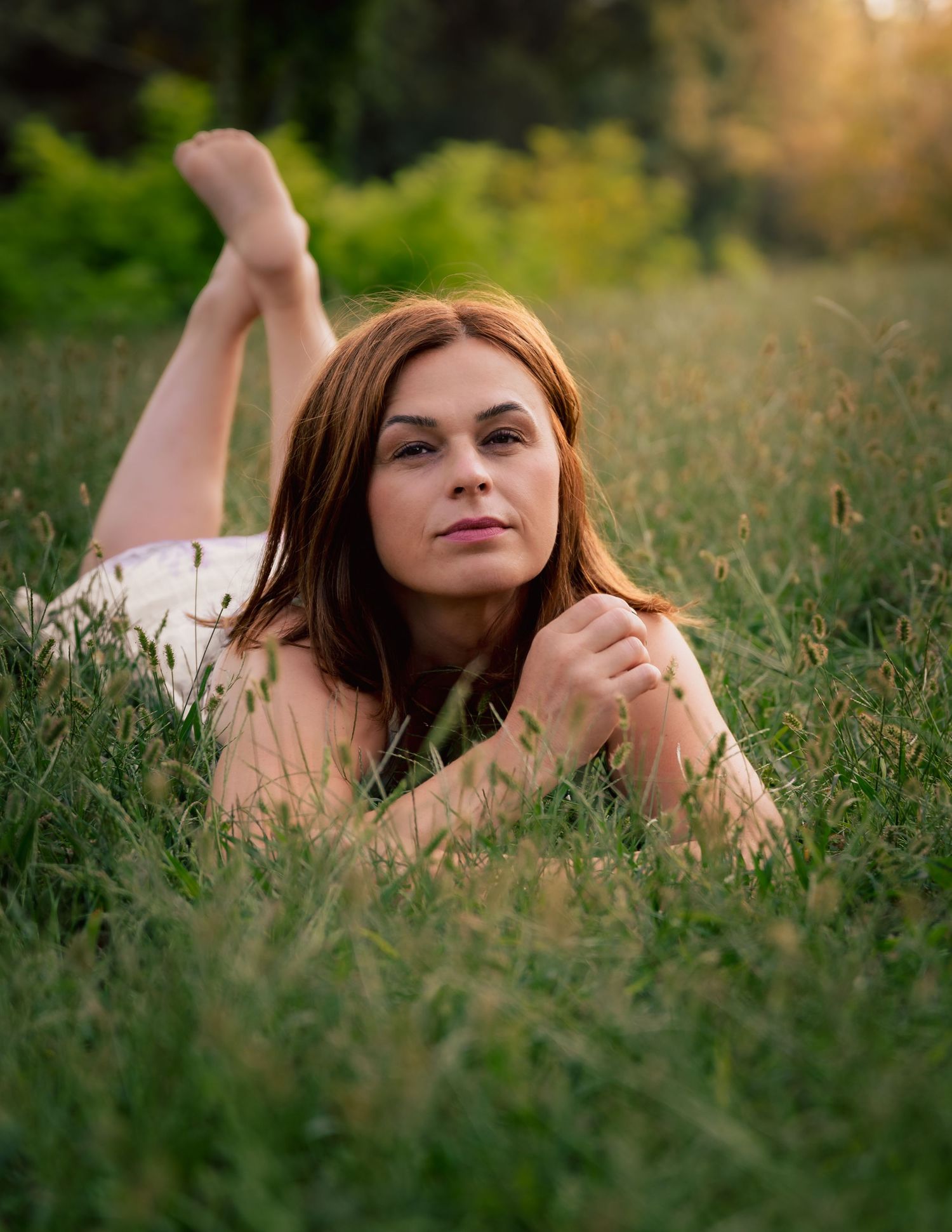 A young woman with auburn hair lies in lush green grass, gazing at camera during golden hour sunset.