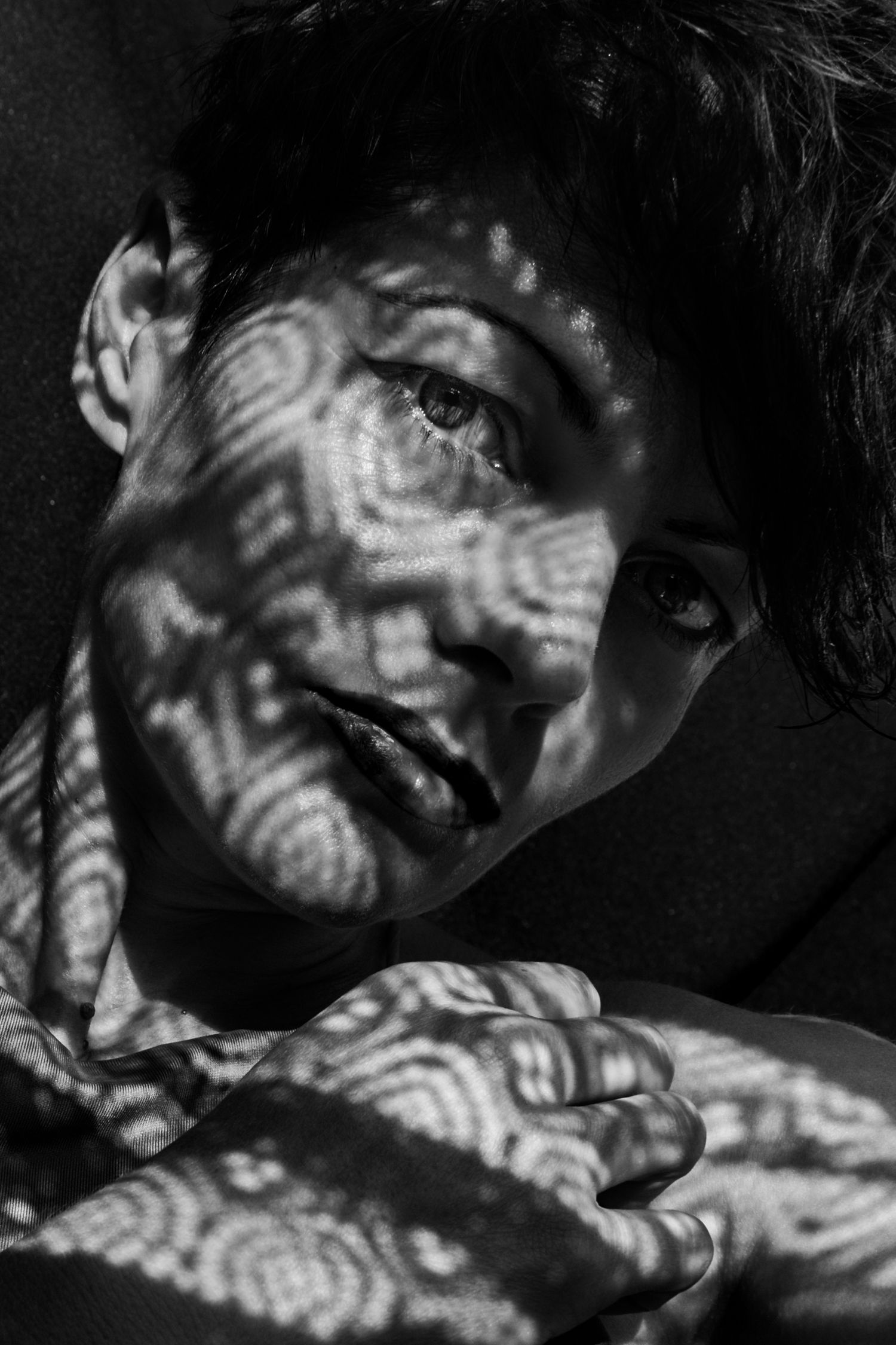 Dramatic black and white portrait with dappled light and shadows creating a moody, film noir aesthetic.