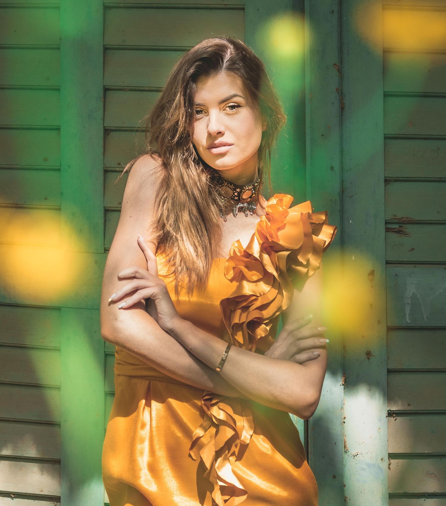 Woman in golden dress holding orange flowers, posing against green wooden wall in warm sunlight.