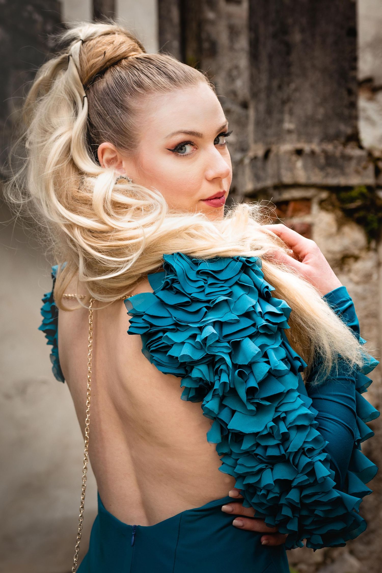 Blonde woman in backless teal ruffled dress poses outdoors, hair flowing, looking over shoulder.