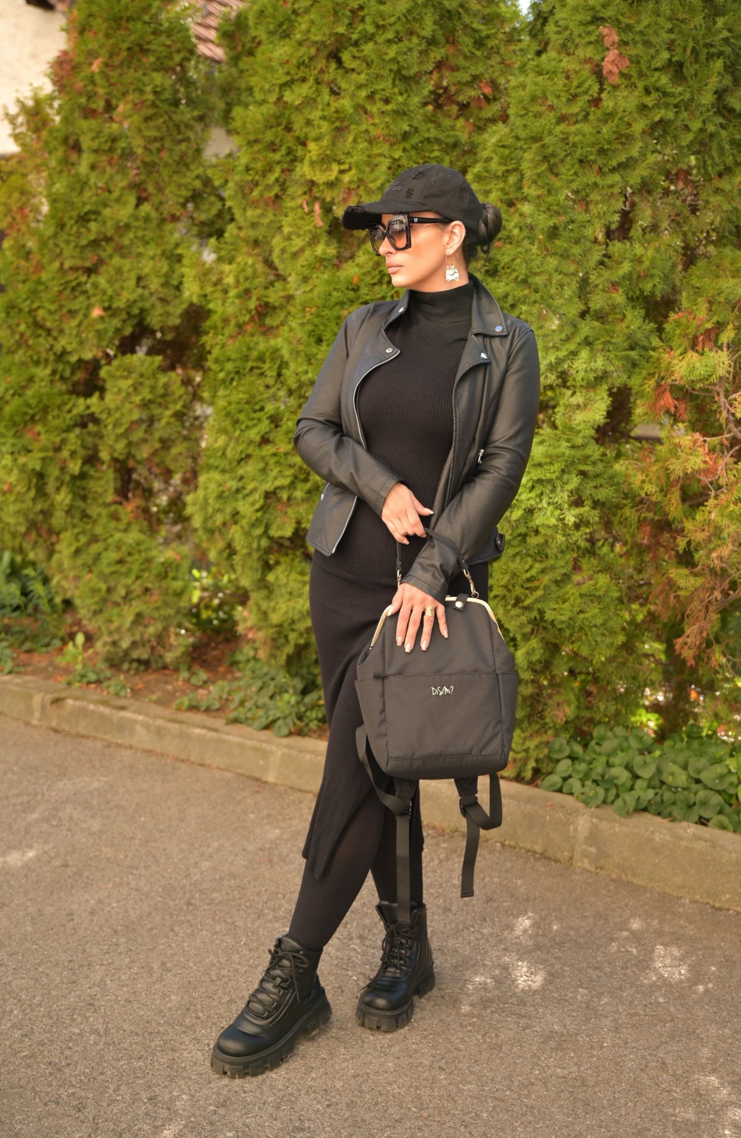 Woman in all-black outfit with leather jacket, cap, sunglasses, combat boots, and gray backpack posing outdoors.