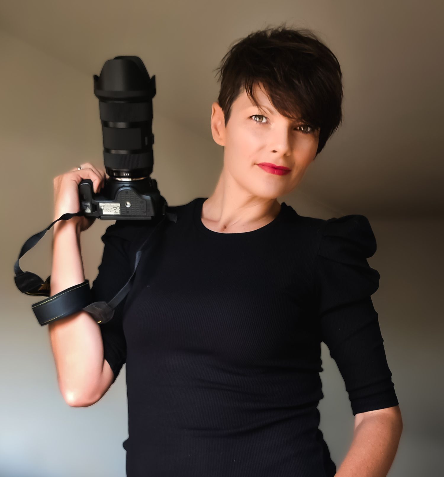 Woman in black dress holding a professional DSLR camera with large lens over her shoulder, posing confidently.