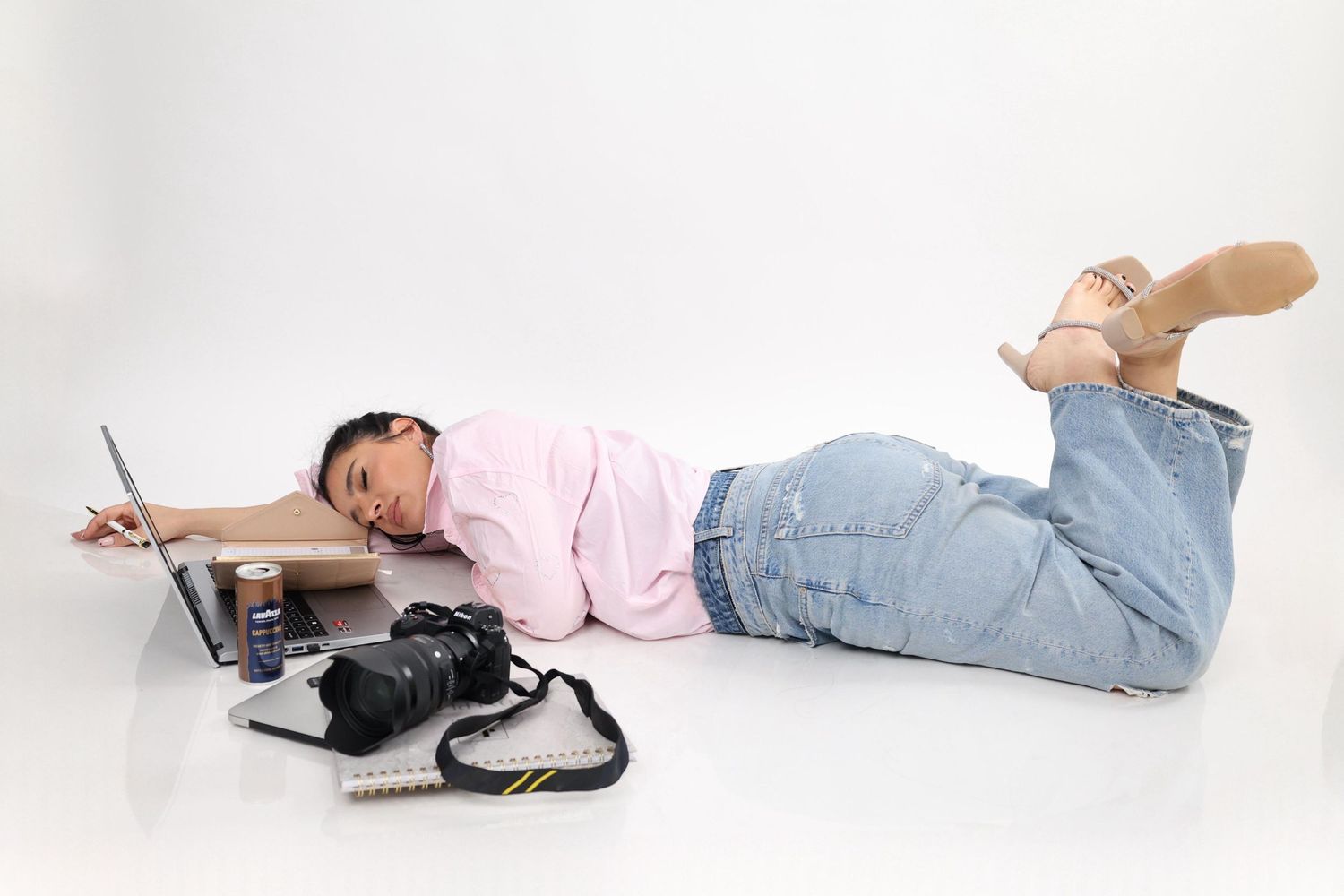 Exhausted female photographer lying on floor with DSLR camera, laptop, and notebook on white background.