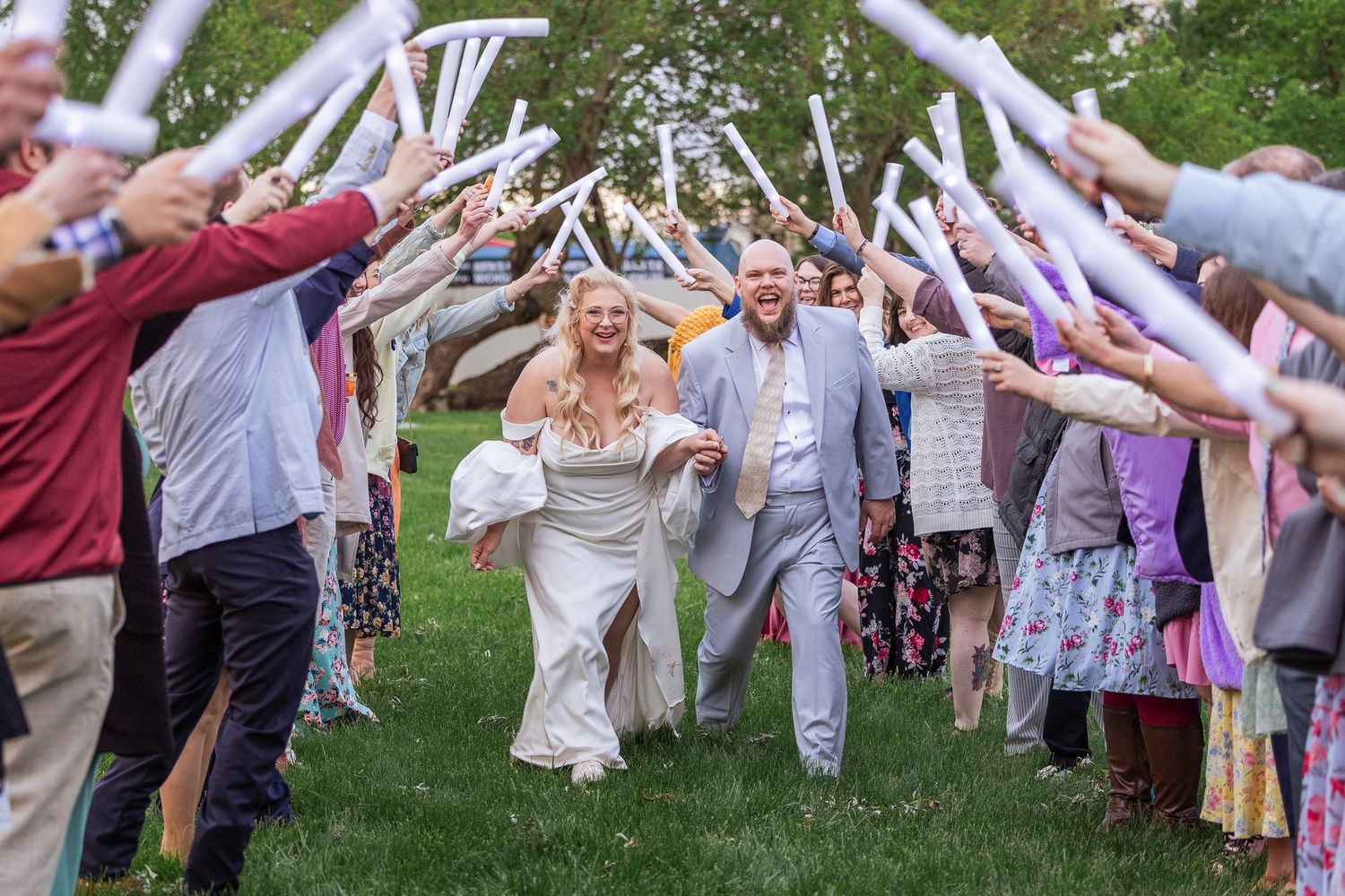 Wedding party guests form honor tunnel with foam glow sticks as newlyweds joyfully run through on grassy lawn at outdoor reception.