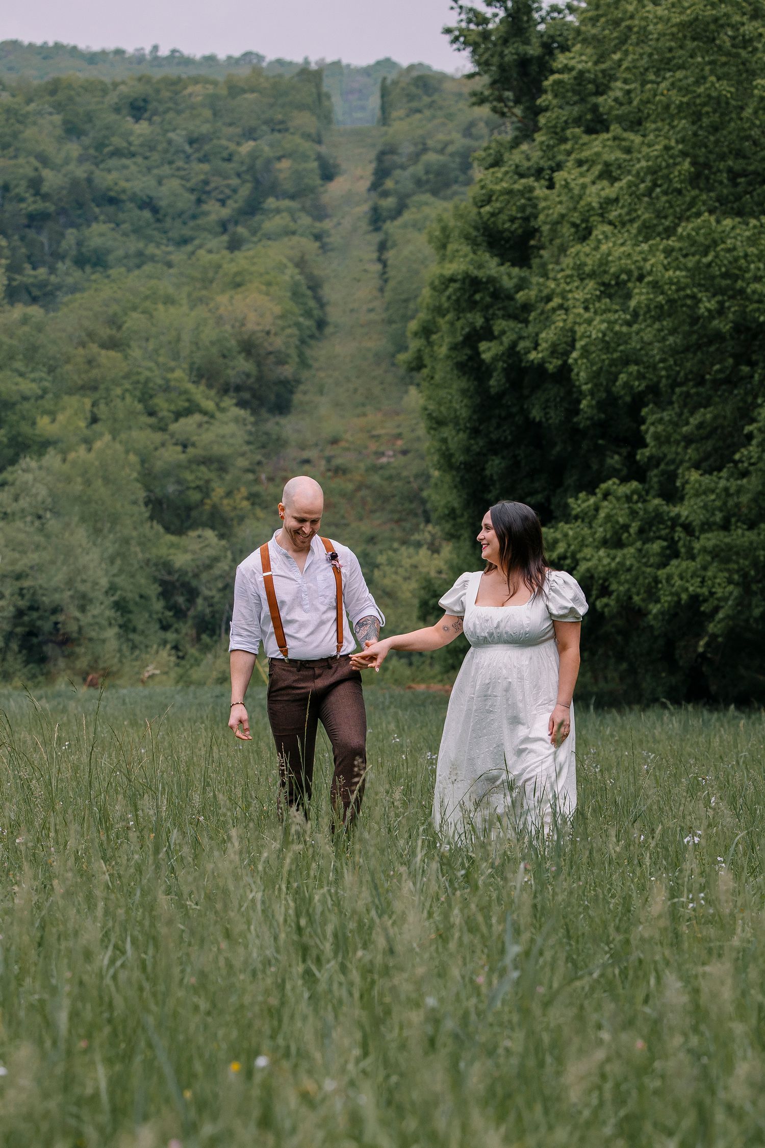 A couple in rustic wedding attire walks through a tall grass meadow with a scenic mountain slope in the background.
