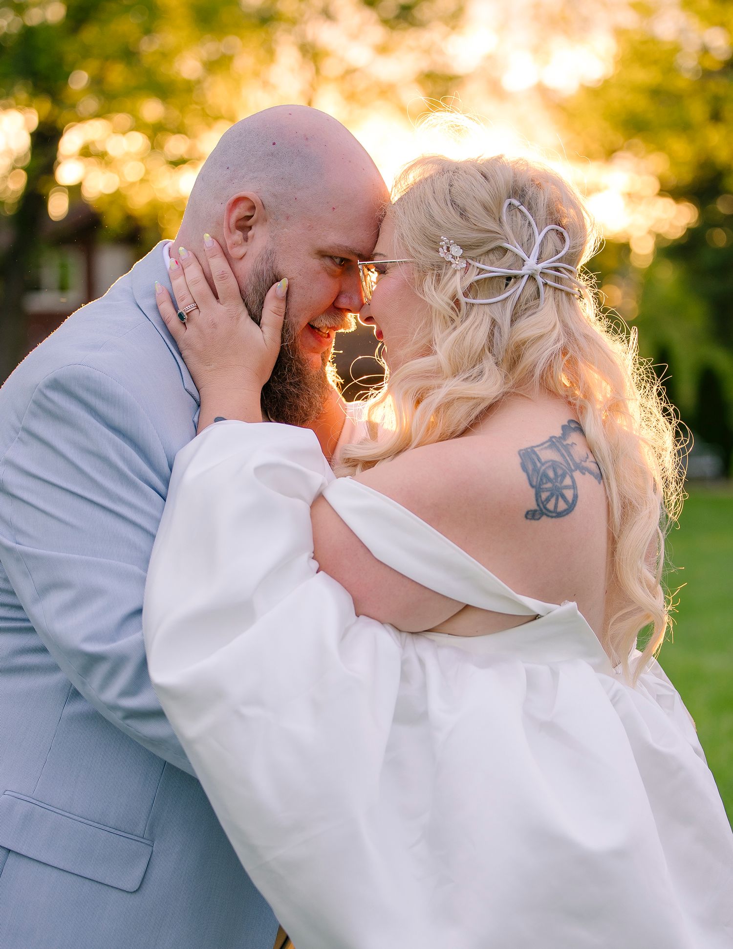 A bald groom with tattoos and a blonde bride in white share an intimate moment outdoors at golden hour.