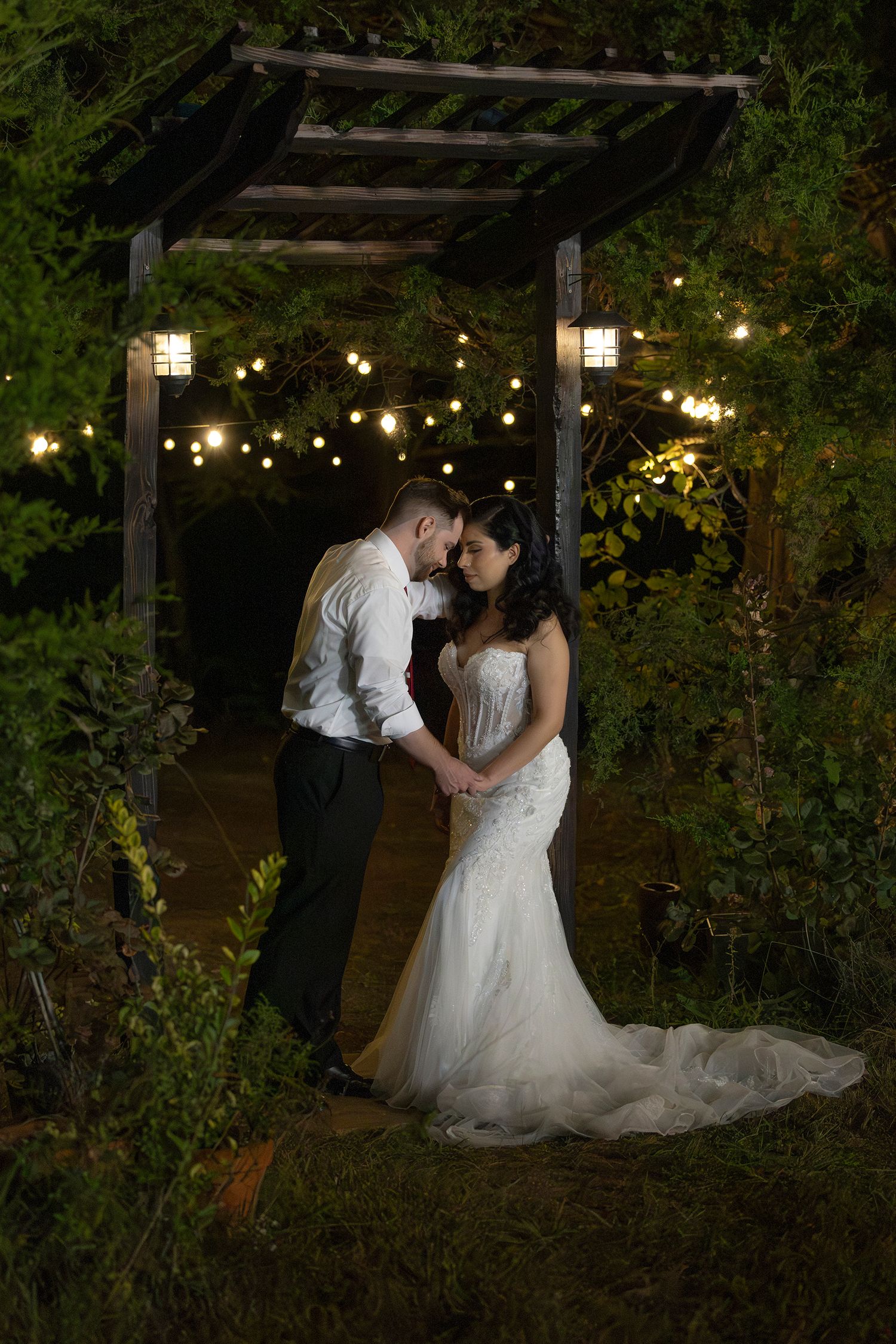 Romantic wedding couple shares an intimate moment under a wooden pergola decorated with twinkling string lights at night.