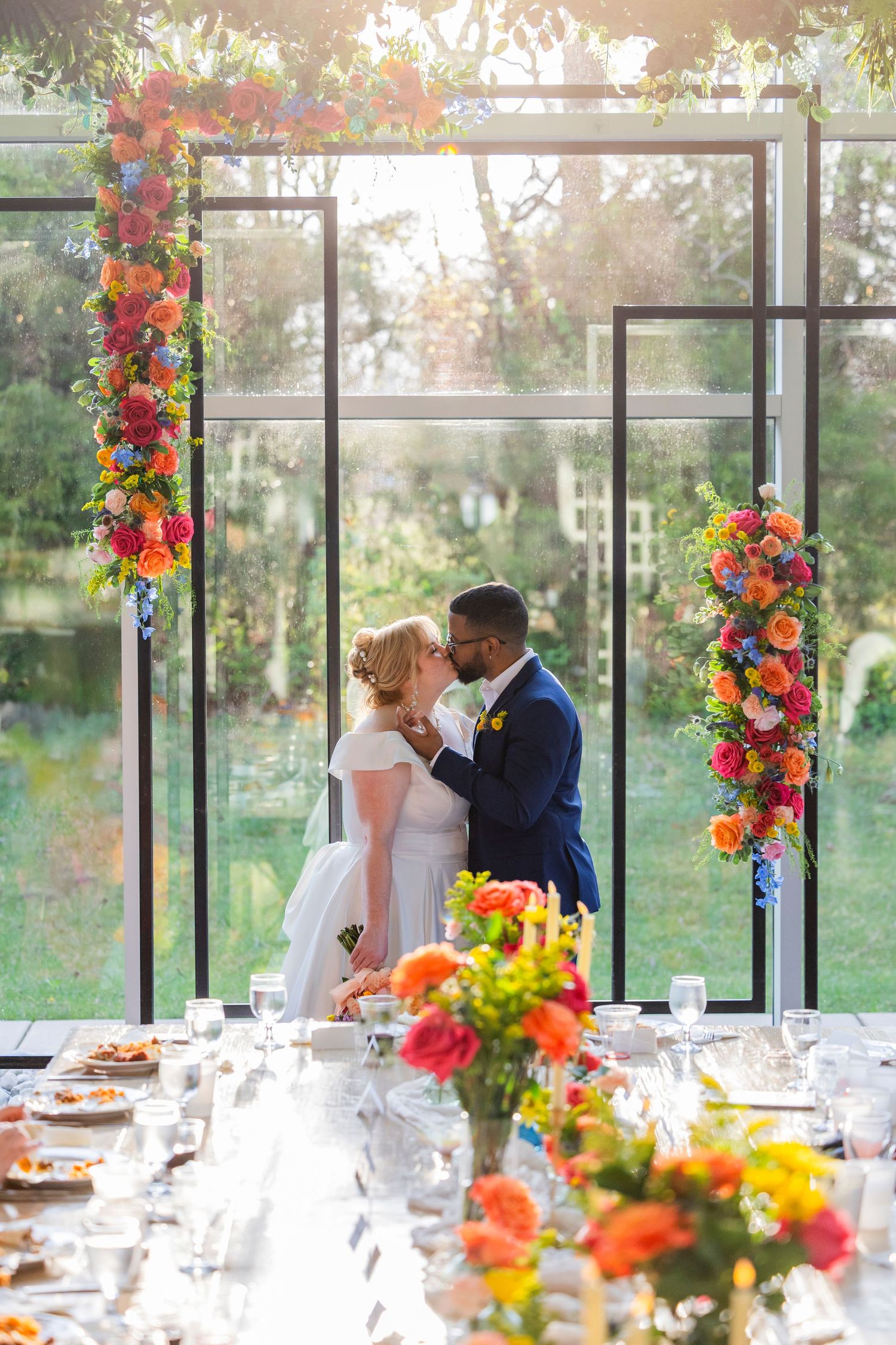 A wedding couple sharing a romantic moment under a modern geometric arch decorated with vibrant orange and red flowers.