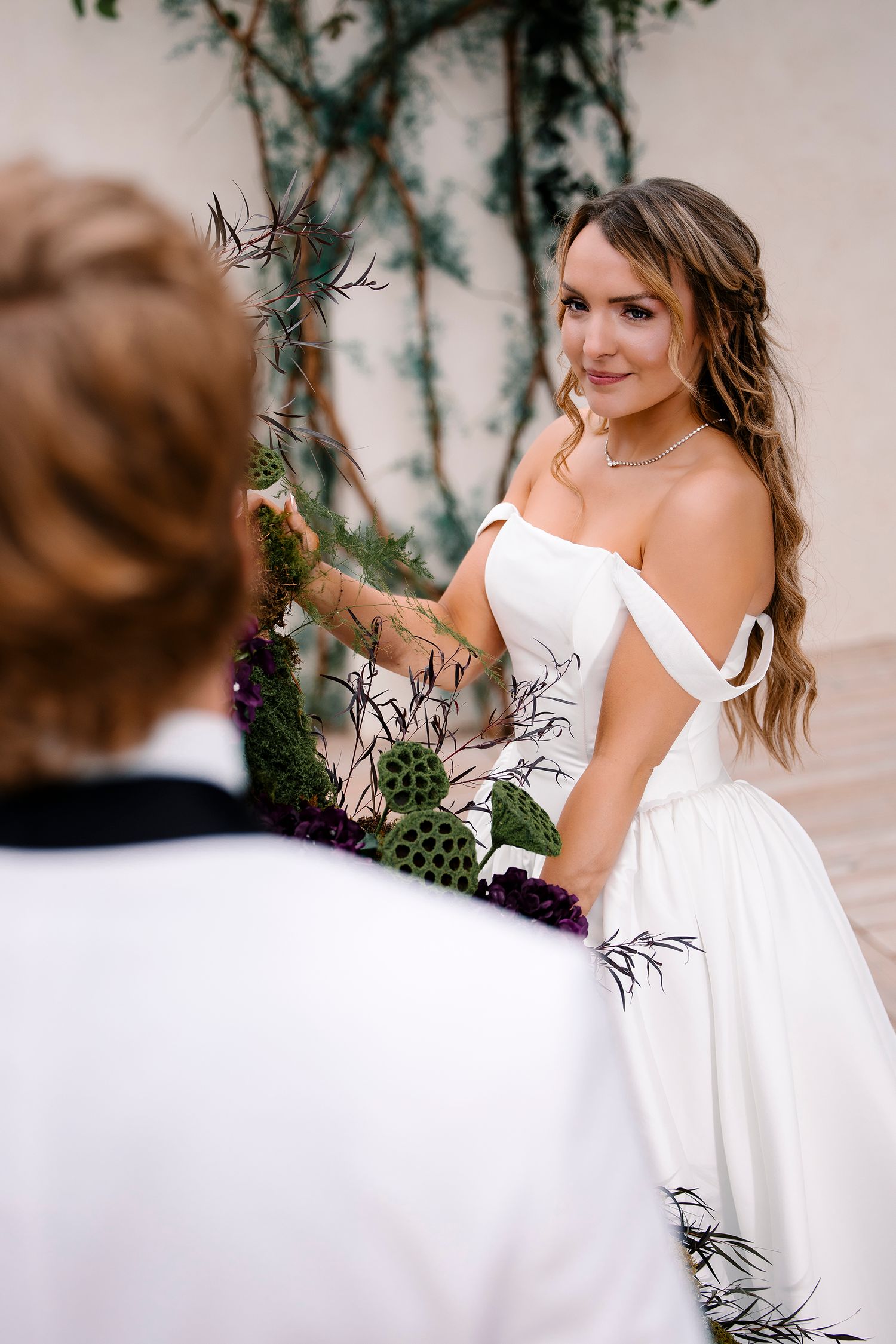 Bride in flowing white dress poses gracefully in garden setting with greenery and floral decor.