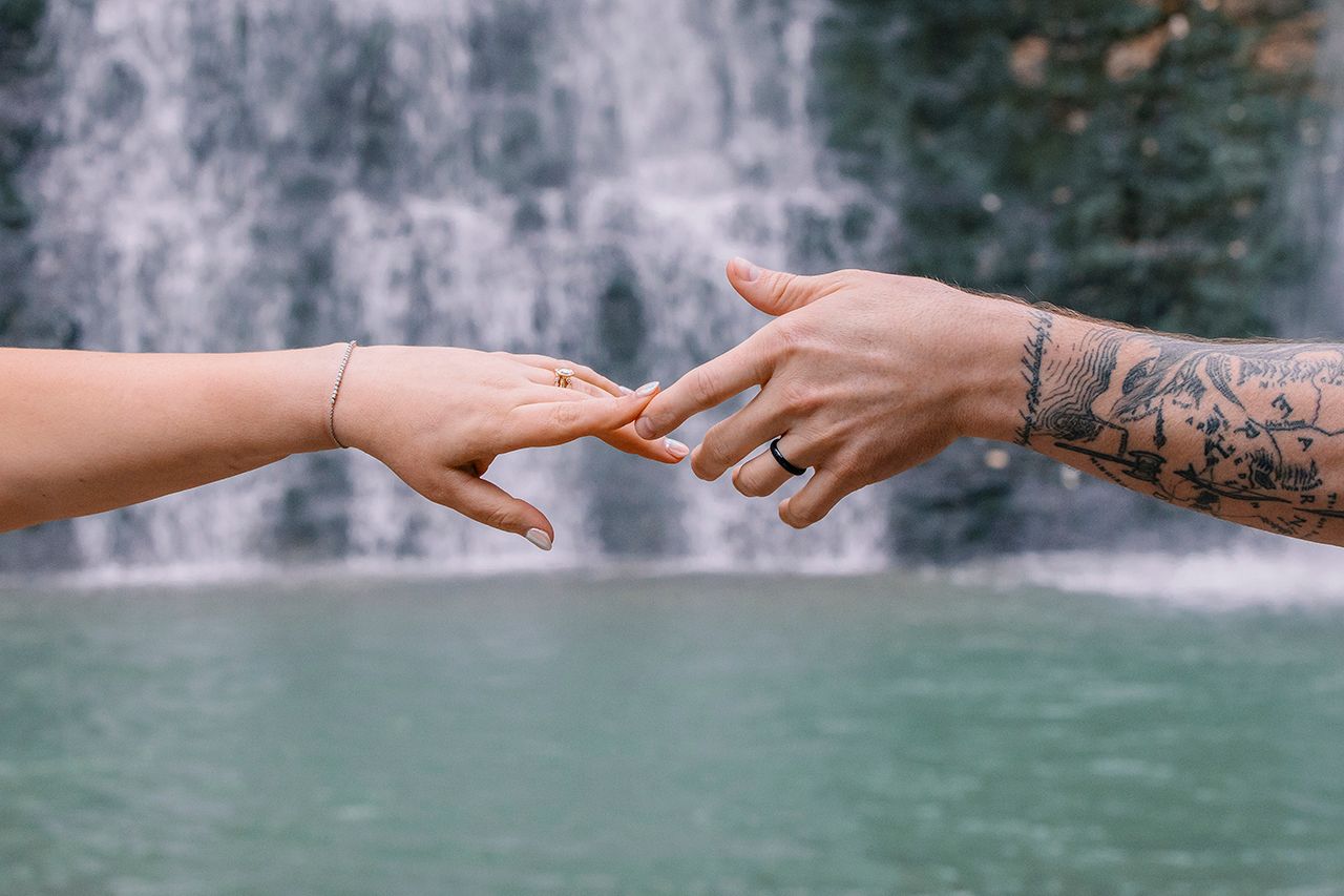 Two hands reaching towards each other with one arm showing detailed tattoo artwork, against a waterfall and turquoise water backdrop.