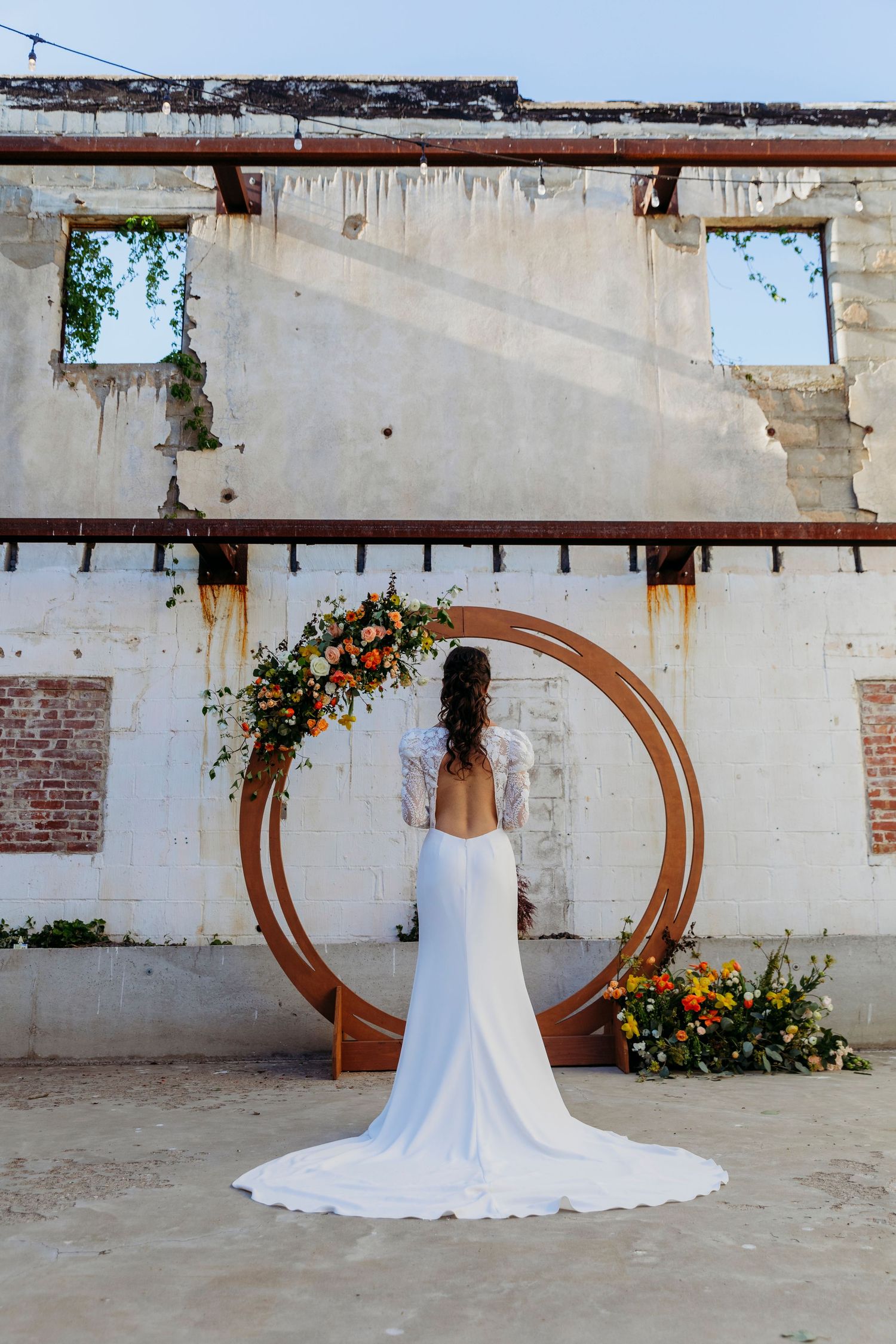 A white wedding dress displayed against a rustic wall with a wooden circular arch decorated with orange flowers.