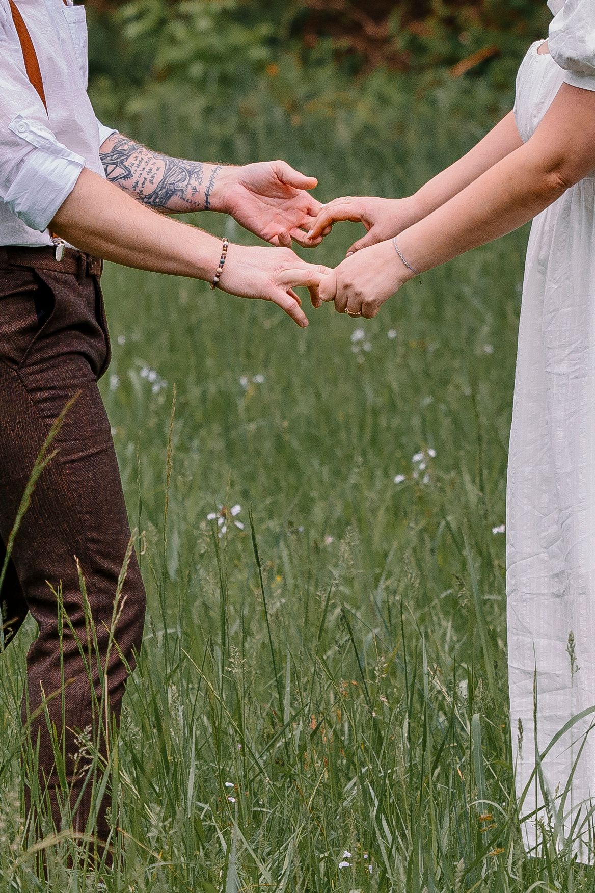 Couple forming a heart shape with their hands in a green meadow, groom with tattoos wearing suspenders.