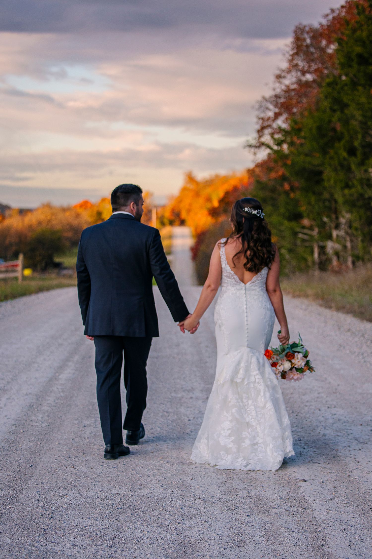 A couple in wedding attire walking down a gravel path at sunset with autumn trees in the background.