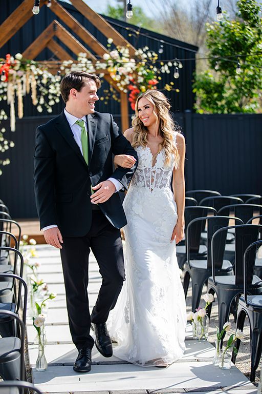 Wedding couple walks down aisle at outdoor ceremony venue with black geometric backdrop and hanging floral decor.