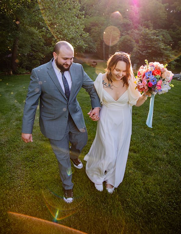 A couple in wedding attire walks hand in hand through a lush garden at sunset with a colorful floral bouquet.