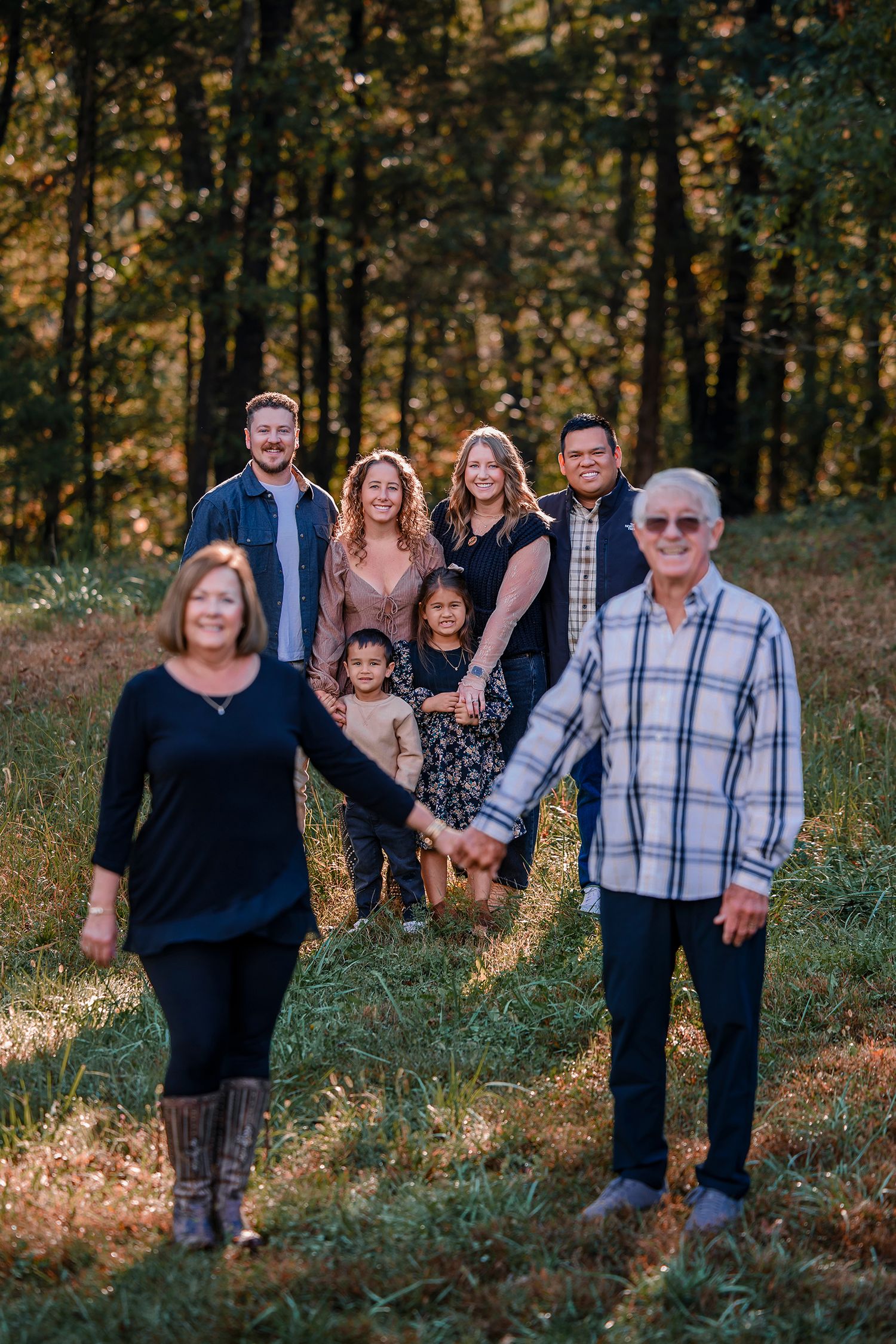 Multi-generational family portrait outdoors in a sunlit forest clearing, smiling and holding hands.