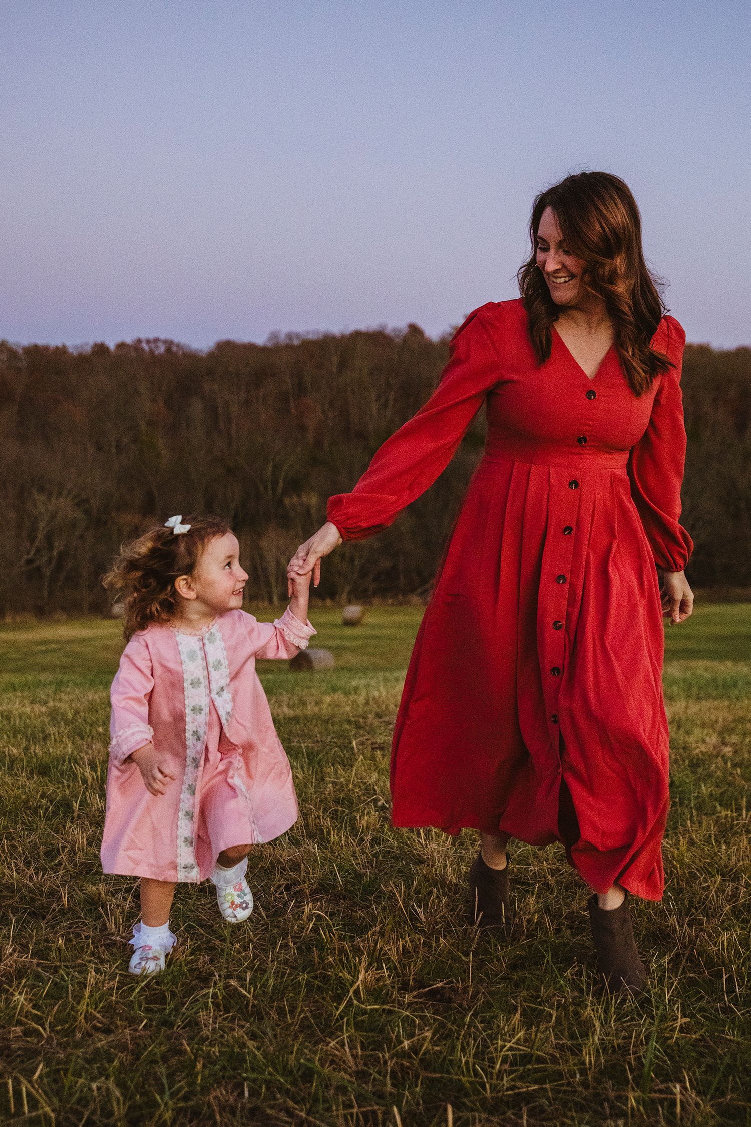 A woman in a red dress holds hands with a toddler girl in pink while walking in a green field at sunset.