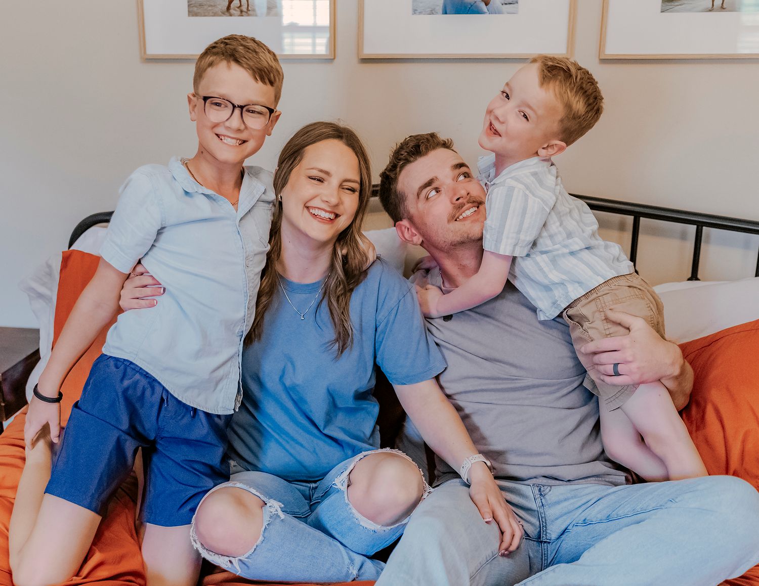 Happy family of four smiling together on a bed, including two young boys and their parents in casual blue outfits.