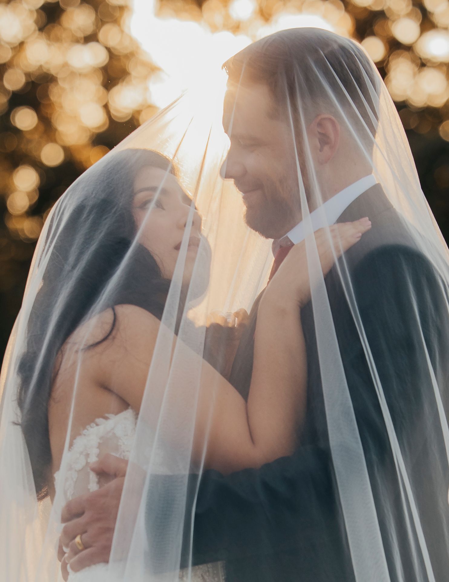 A romantic wedding couple shares an intimate moment under a flowing white veil with soft golden bokeh in the background.