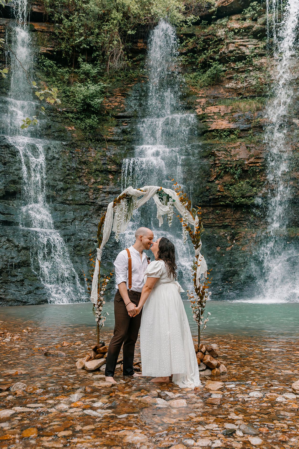 A rustic wedding arch decorated with floral arrangements stands before three cascading waterfalls in a scenic forest setting.