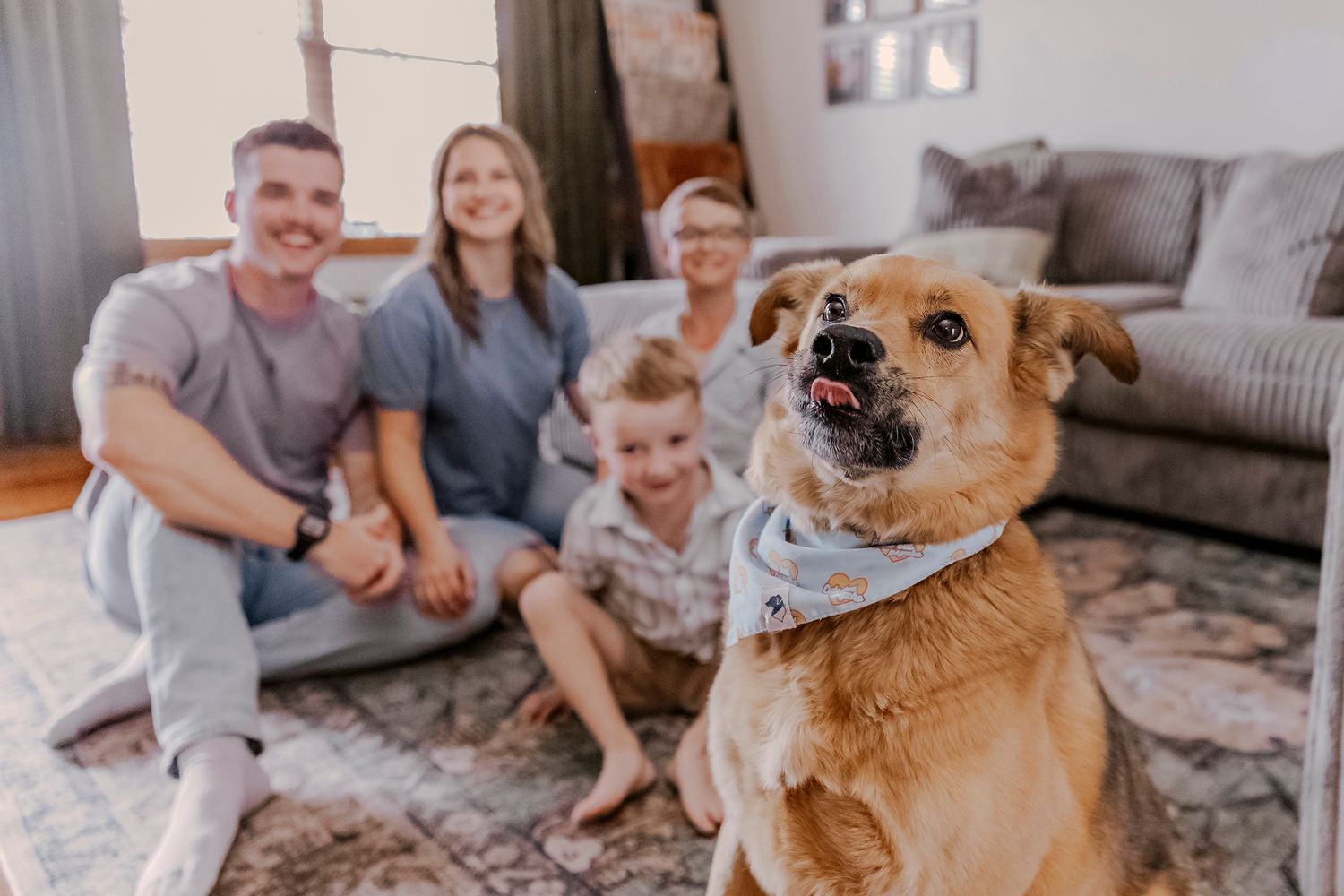 Happy family with dog wearing bandana sitting together on living room floor with couch in background.