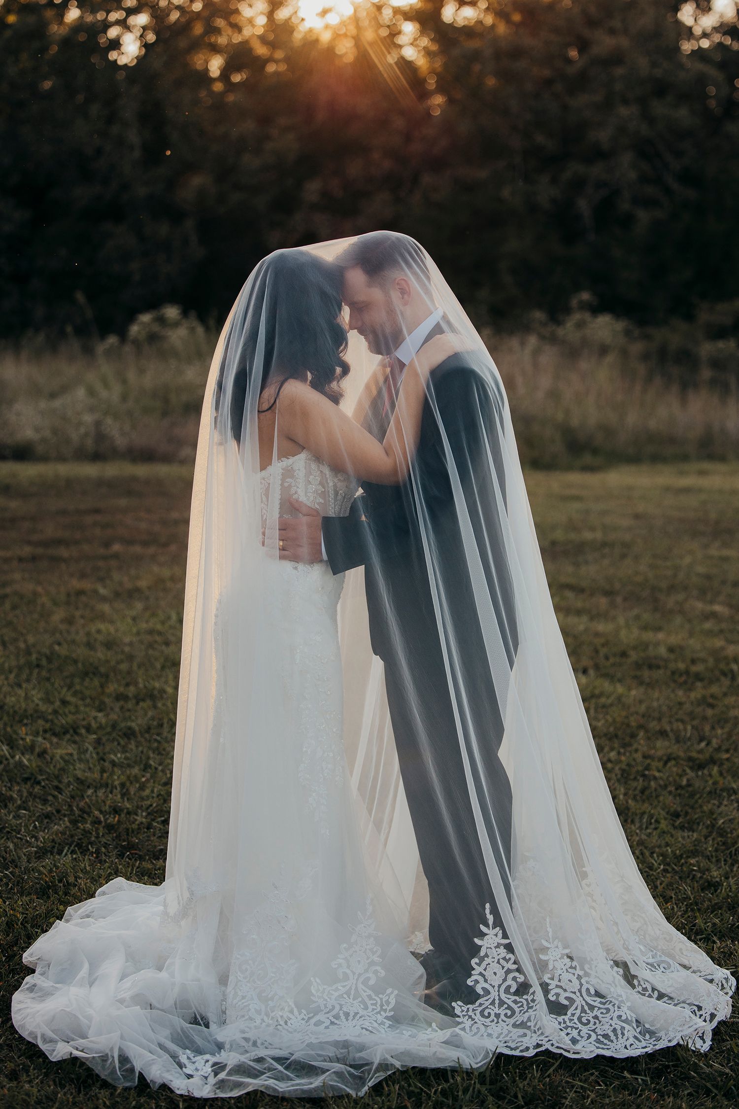 Bride and groom share intimate moment under flowing lace veil at golden hour sunset in lush garden setting.