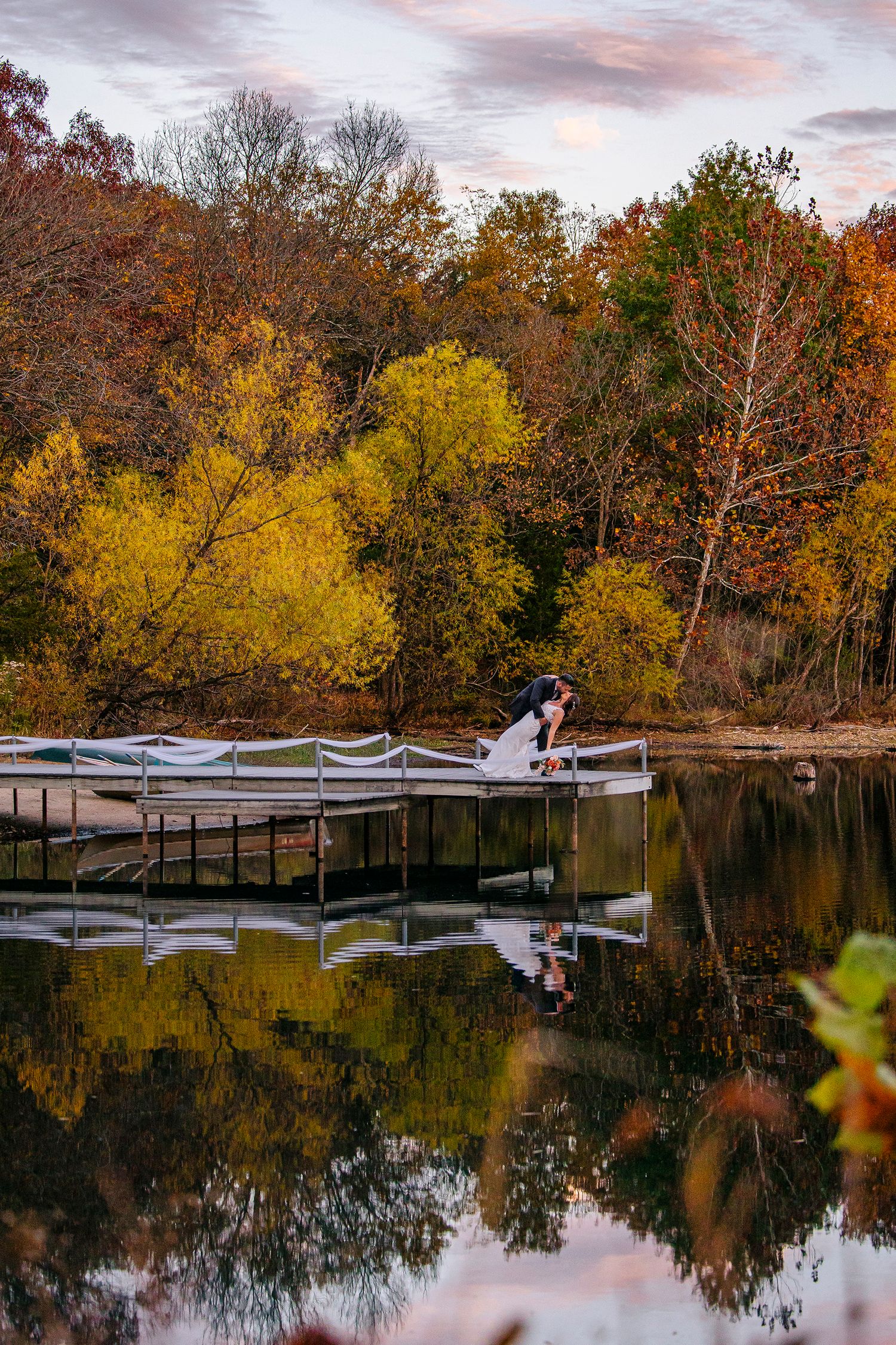 Couple sitting on wooden dock surrounded by autumn trees reflecting in calm lake water at sunset.