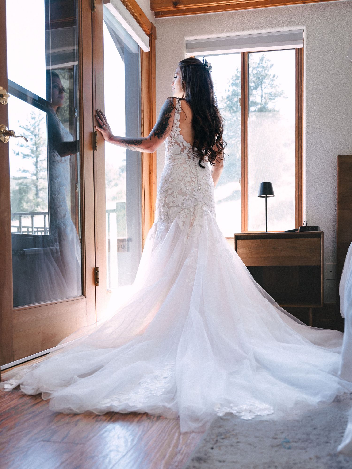 A stunning lace wedding dress with a long flowing train is silhouetted against bright natural light from tall windows.