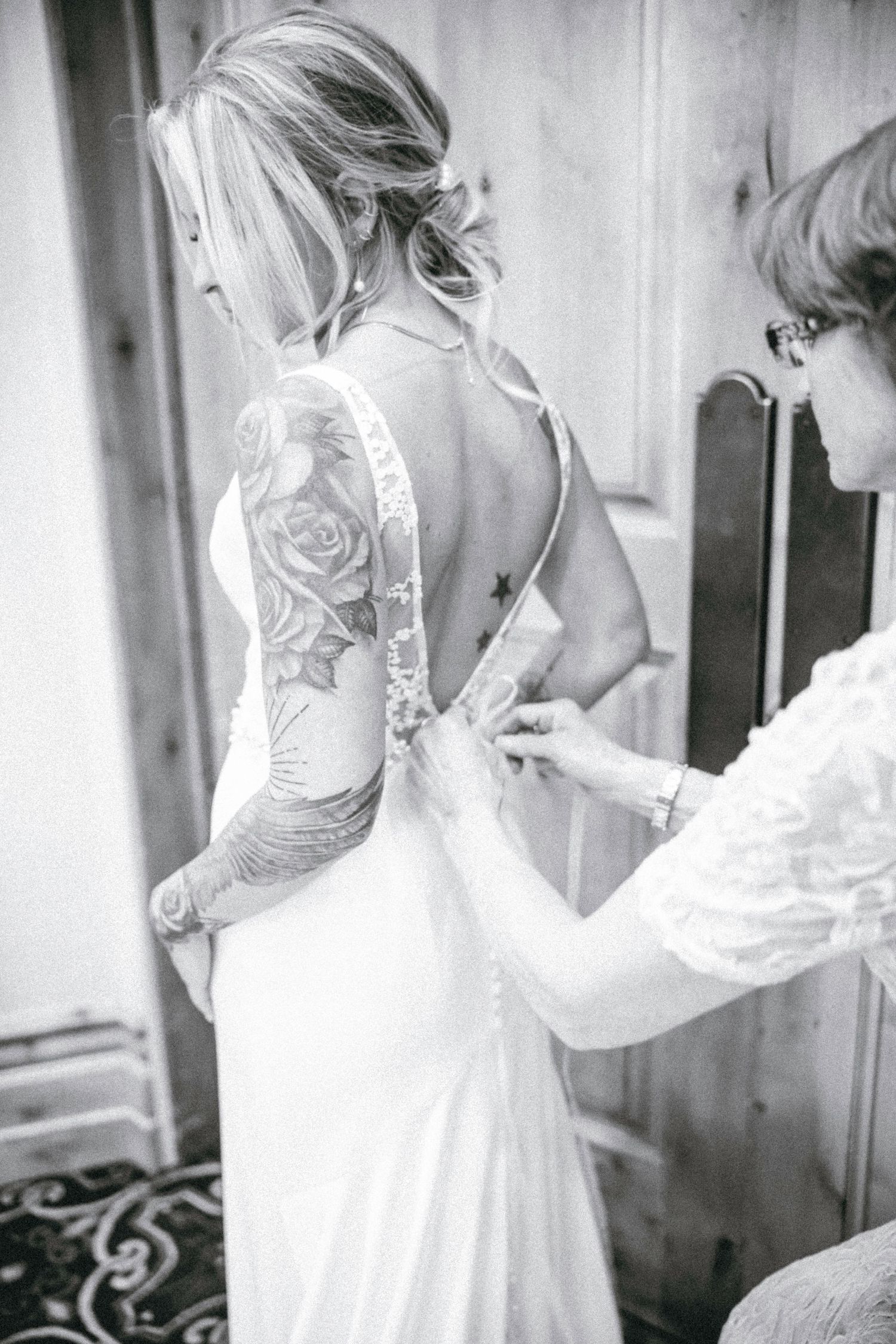 Black and white photo showing a bride in a backless wedding dress with rose tattoo sleeves being buttoned up.