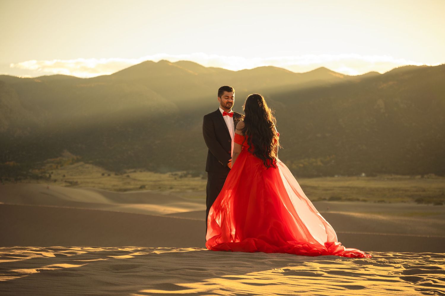 A couple stands on sand dunes at sunset with mountains in background as red dress flows in the wind.
