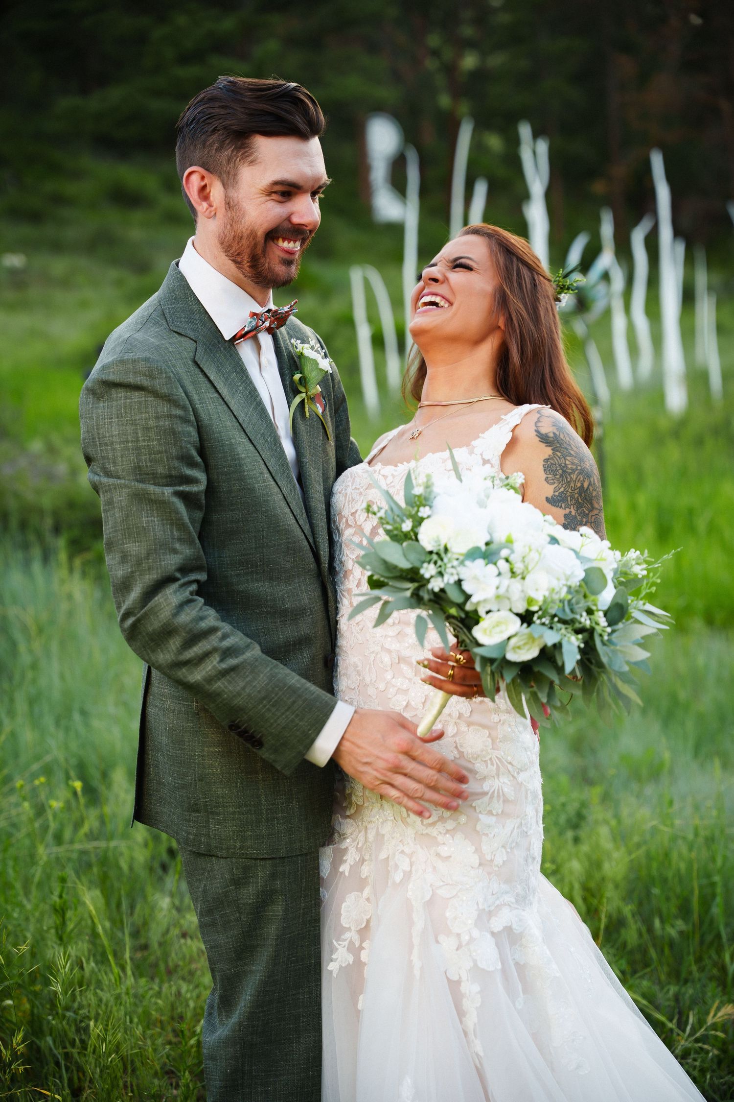 A joyful wedding couple embraces while laughing in a grassy meadow with white fence posts in the background.