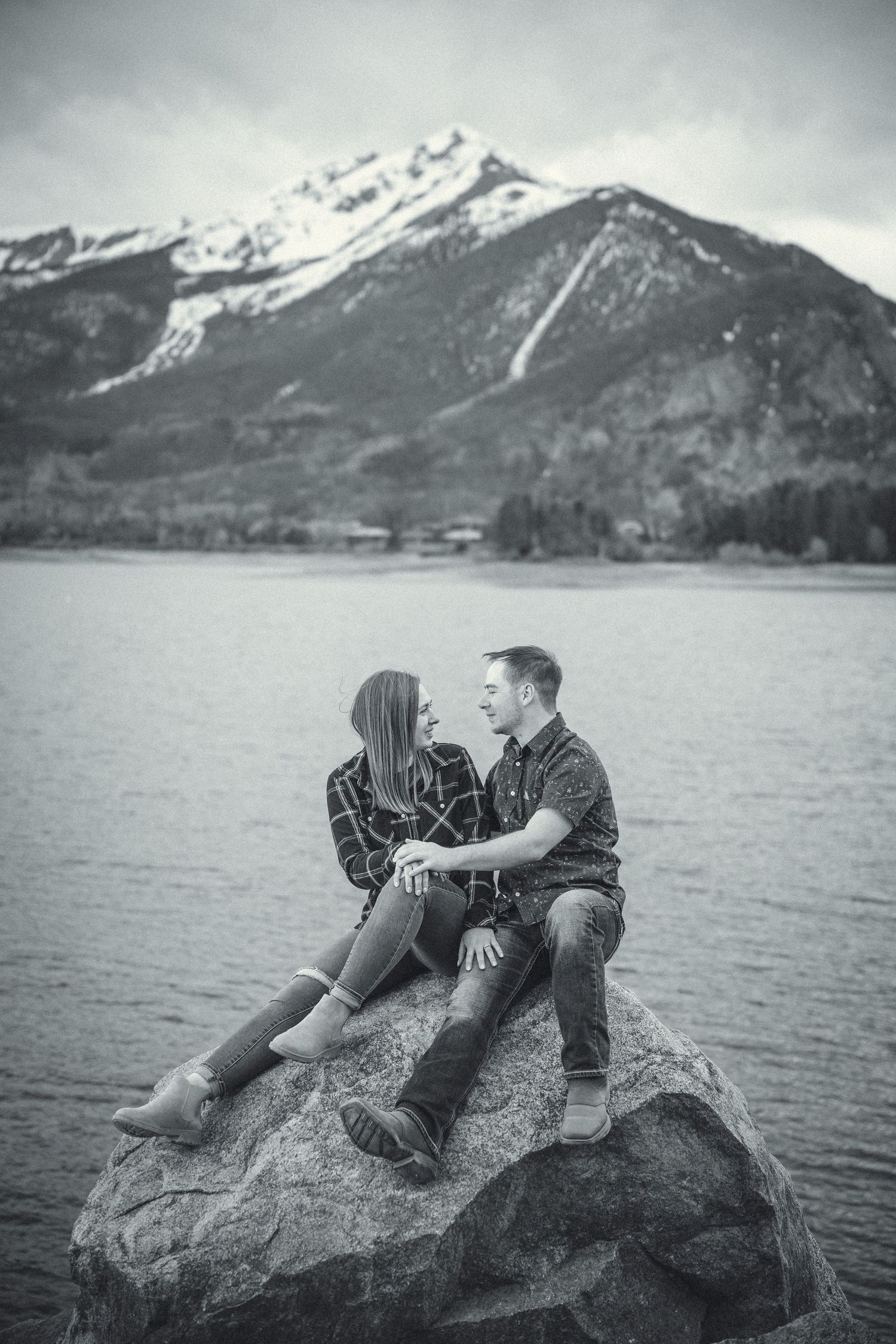 A romantic couple lounges on large rocks by a scenic mountain lake in black and white photography.