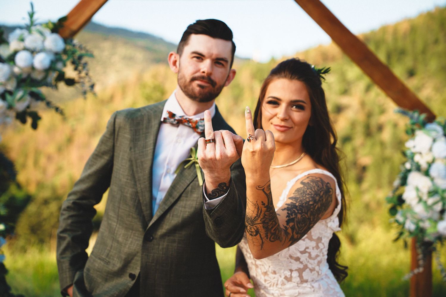 Wedding couple embraces under rustic wooden arch decorated with floral arrangements in golden outdoor setting.