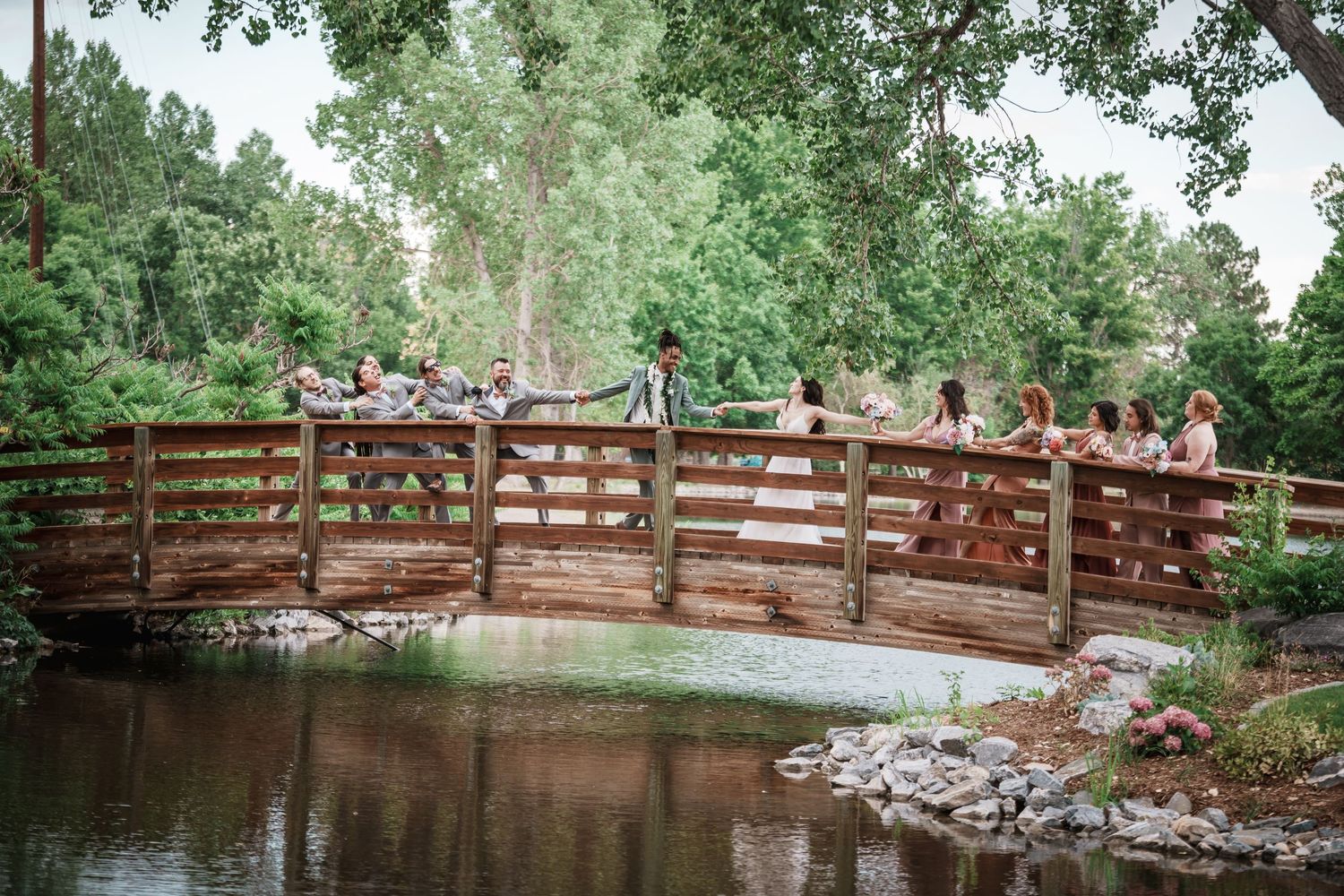 Wedding party gathers on a wooden footbridge over a tranquil pond surrounded by lush green trees in a park setting.