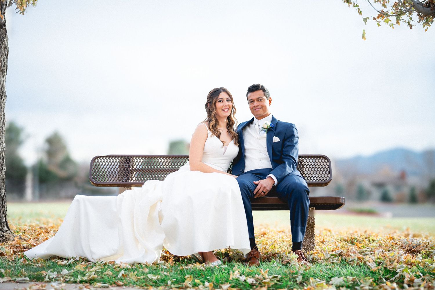 A newlywed couple sits on a rustic bench surrounded by autumn leaves in an outdoor setting with mountains in the distance.