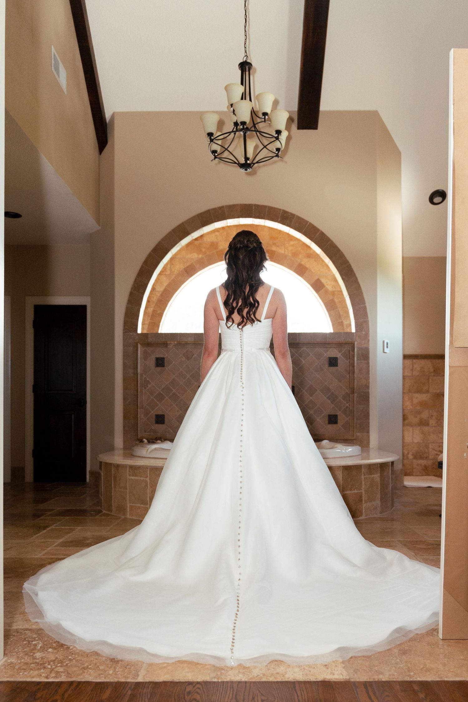 A bride in an elegant white wedding gown with a sweeping train stands before an arched doorway under a chandelier.