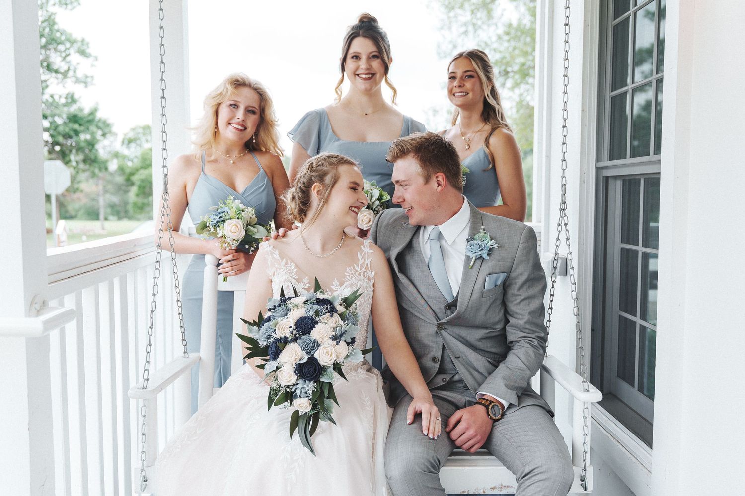 Wedding party laughs together while posing on an elegant white porch in light summer attire.