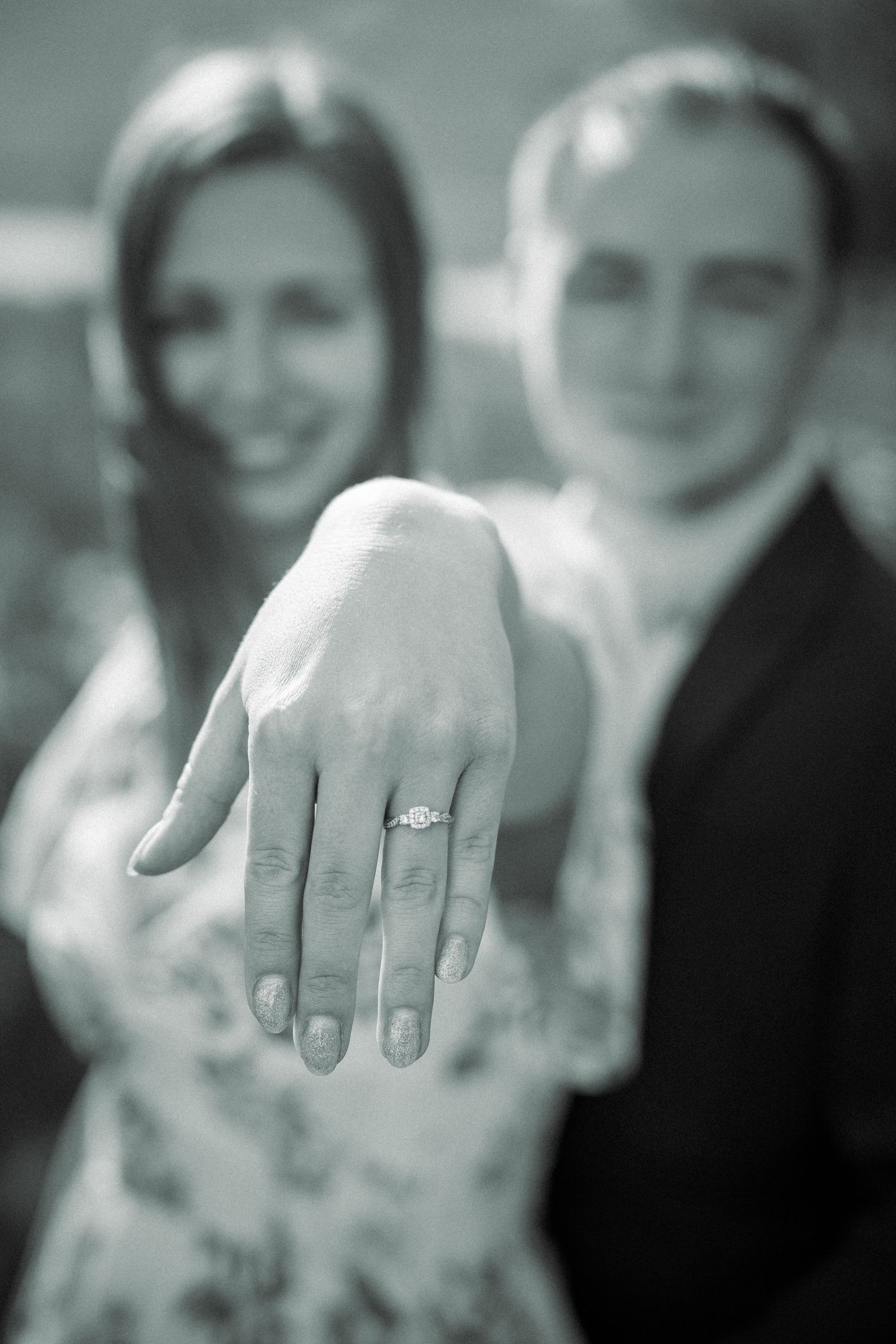 Close-up of a newly engaged hand showing off diamond engagement ring with couple blurred in background.