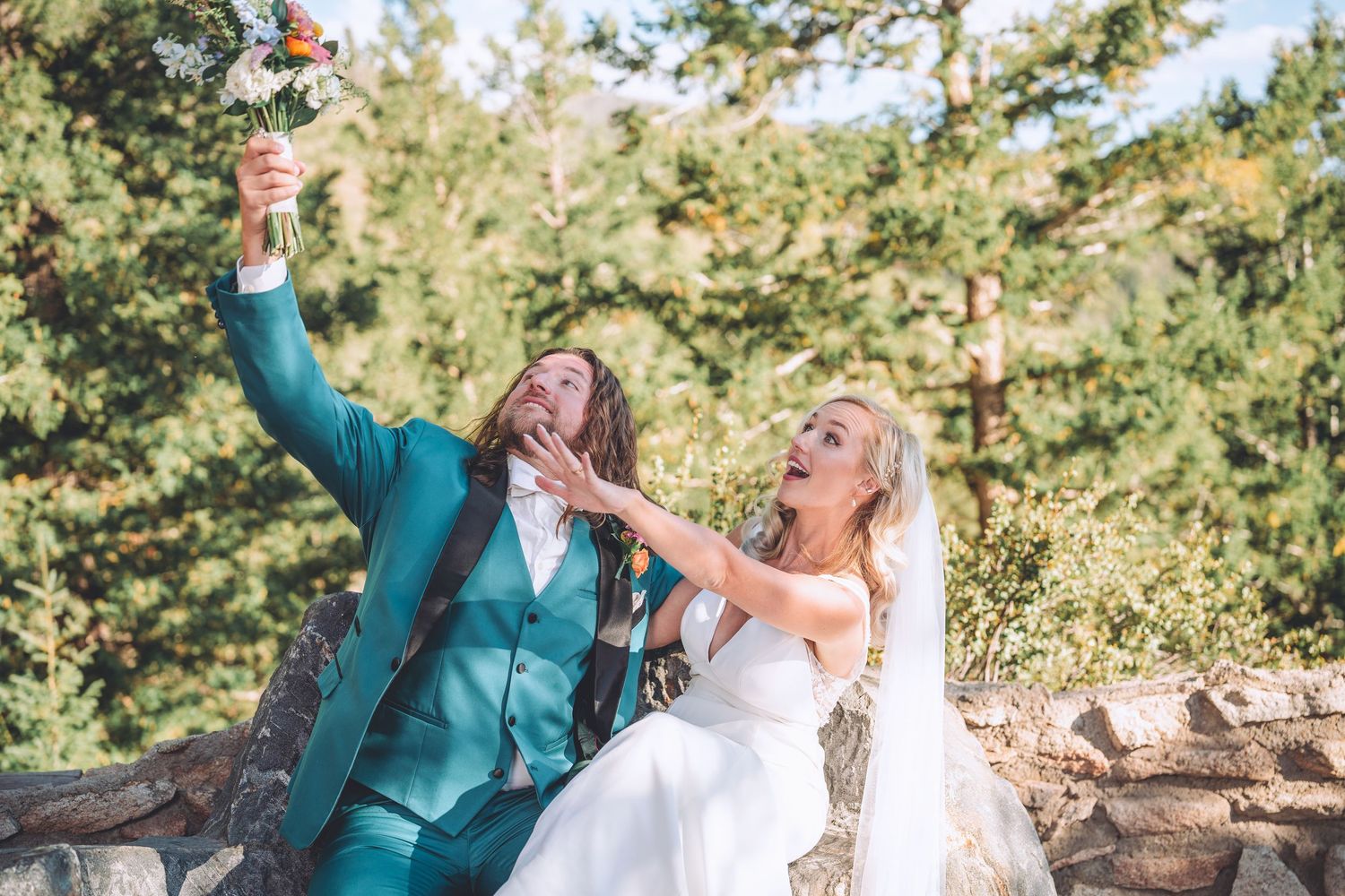 A couple in wedding attire laugh and celebrate while sitting outdoors in a natural setting.