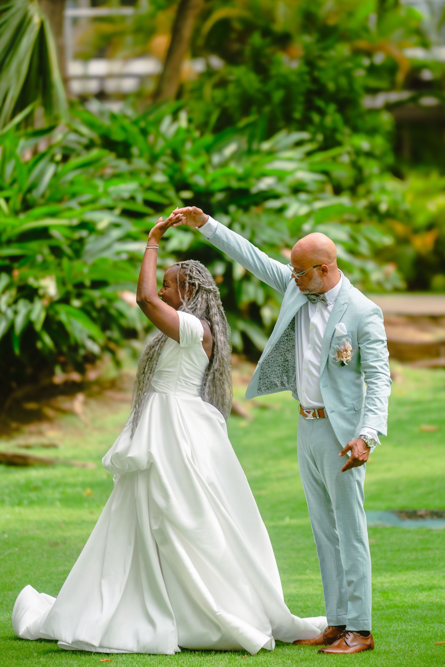 A couple in wedding attire dancing together on a lush green lawn surrounded by tropical plants.