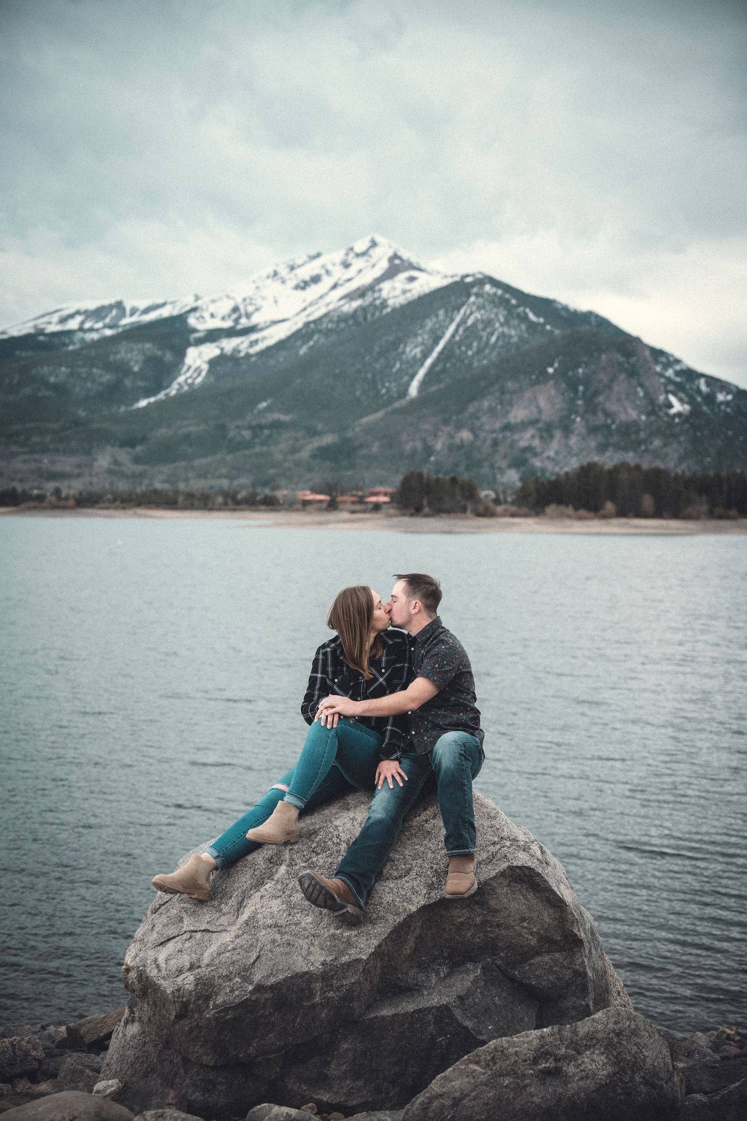 A couple sits together on a large boulder by a mountain lake with snow-capped peaks in the background.