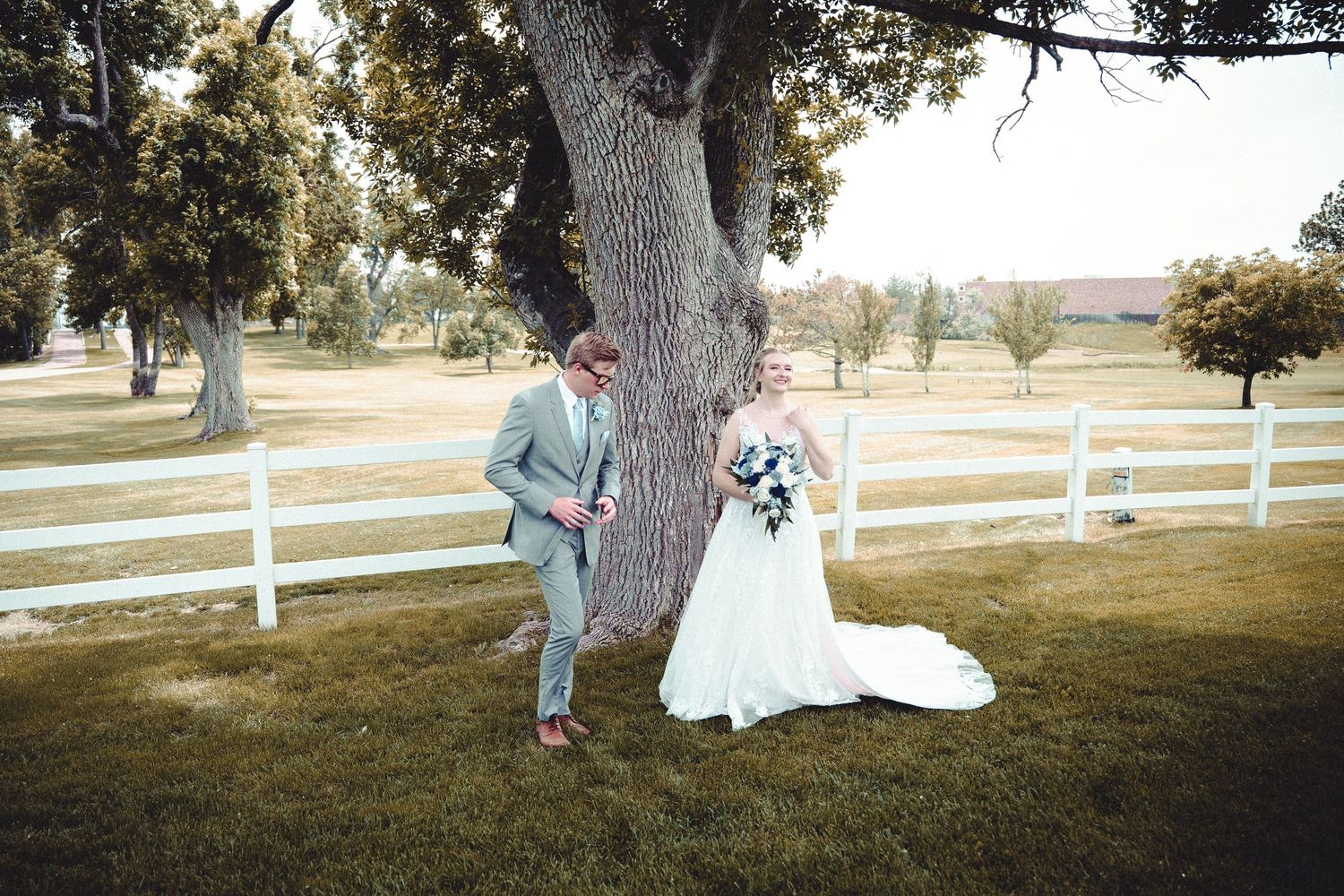 Two people in wedding attire stand together under a large tree in a scenic outdoor setting.